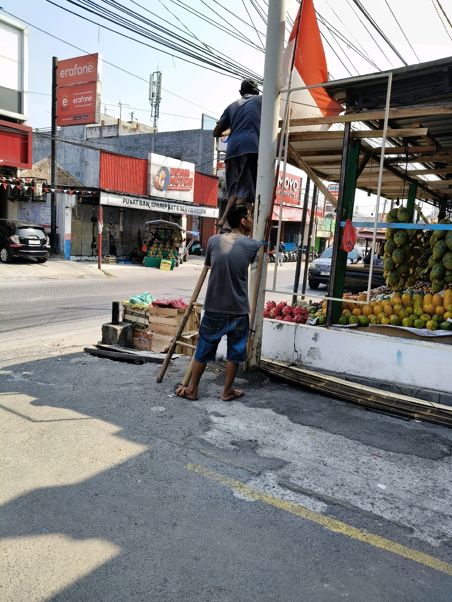 The image depicts a street scene in Bekasi, Indonesia, with two individuals interacting with a flagpole. One man, wearing a dark blue t-shirt and denim shorts, is standing on a wooden ladder, appearing to adjust a red and white Indonesian flag attached to the pole. Another man, dressed in a grey t-shirt and dark shorts, stands at the base of the ladder, looking upwards. The background reveals commercial establishments, including a building with red accents and signage for "e-rafone." Further down the street, a fruit stand is visible, displaying an abundance of coconuts, mangoes, and other produce. The environment is characterized by a paved road, overhead power lines, and clear daylight conditions.