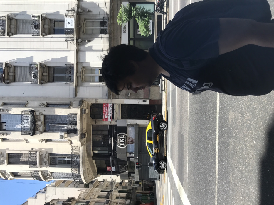 A street scene in Buenos Aires is captured with a sideways perspective. A black and yellow taxi is parked on the street, adjacent to a building with ornate balconies. A man stands partially in the frame, occupying the upper right corner, obscuring the sky. A sign reads "ICBC" on one of the buildings. Light and shadows play across the scene.