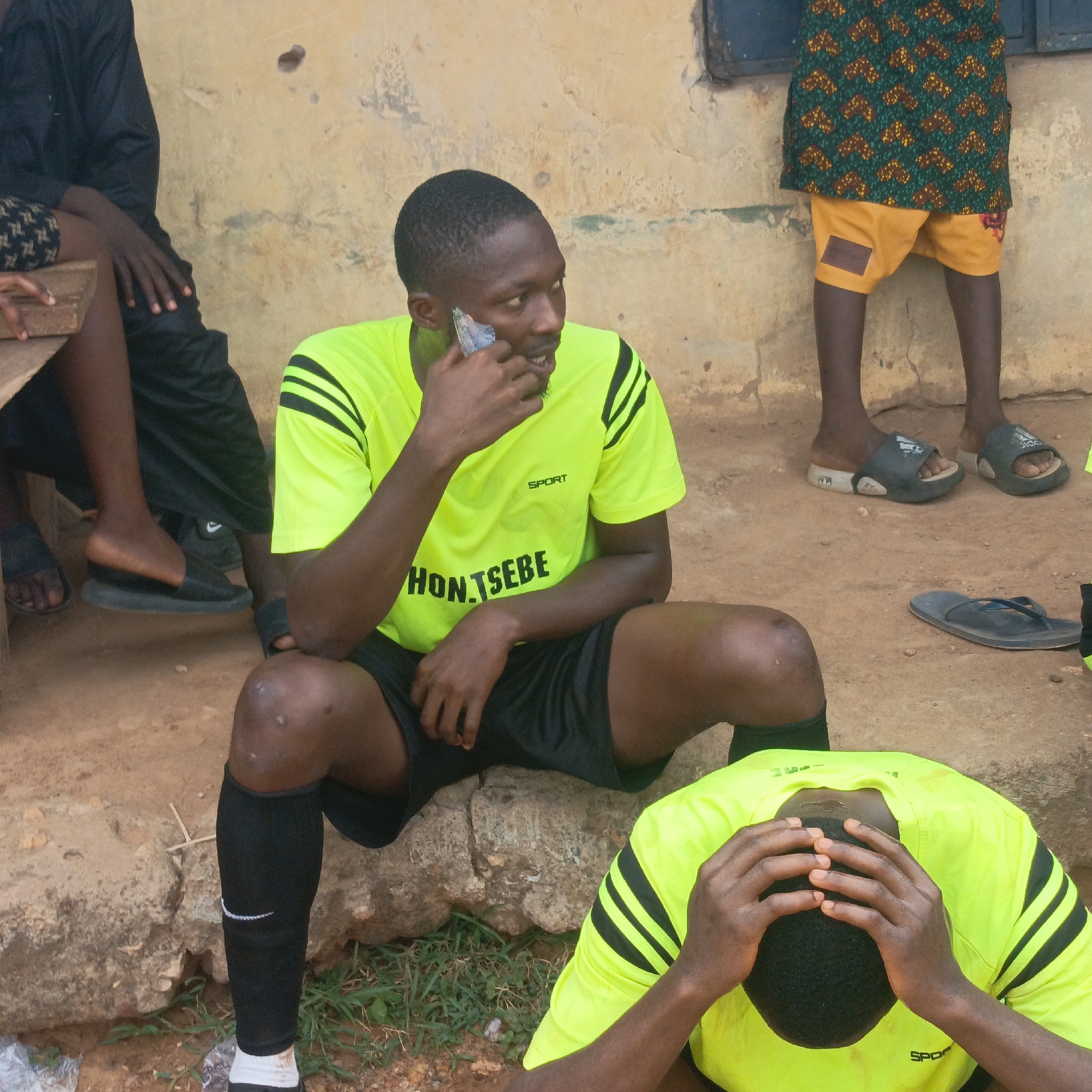 This outdoor photograph, taken in Akwanga, Nigeria, captures a candid moment among several individuals, likely during a break from a sporting activity. In the foreground, a dark-skinned man is seated on a low stone or concrete ledge. He wears a bright neon yellow short-sleeved sports jersey with black stripes on the shoulders, black shorts, and black knee-high socks featuring a white Nike swoosh. He is engaged in a phone call, holding a silver cell phone to his ear, with a thoughtful or slightly concerned expression on his face. Visible text on his shirt includes "SPORT" and "HON. TSEBE".
To the right and lower in the foreground, another individual, similarly dressed in a neon yellow sports jersey, has their head bowed and hands clasped over their head, conveying a strong sense of dejection, disappointment, or frustration. In the left background, a person in dark clothing sits on a wooden bench, with only their lower body visible. On the right side of the frame, a child or shorter person stands, dressed in a patterned green, brown, and yellow top over yellow shorts, wearing dark sandals.
The setting is a somewhat rustic outdoor area. The background features a weathered, light brown or yellowish building wall with a dark window frame visible in the upper right. The ground is dusty earth with sparse patches of green weeds. The overall scene suggests a warm climate and a casual, possibly non-affluent, environment. The sports attire and the clear emotion of dejection suggest the photograph was taken after a game or event where the outcome was not favorable. The time of day appears to be daylight.