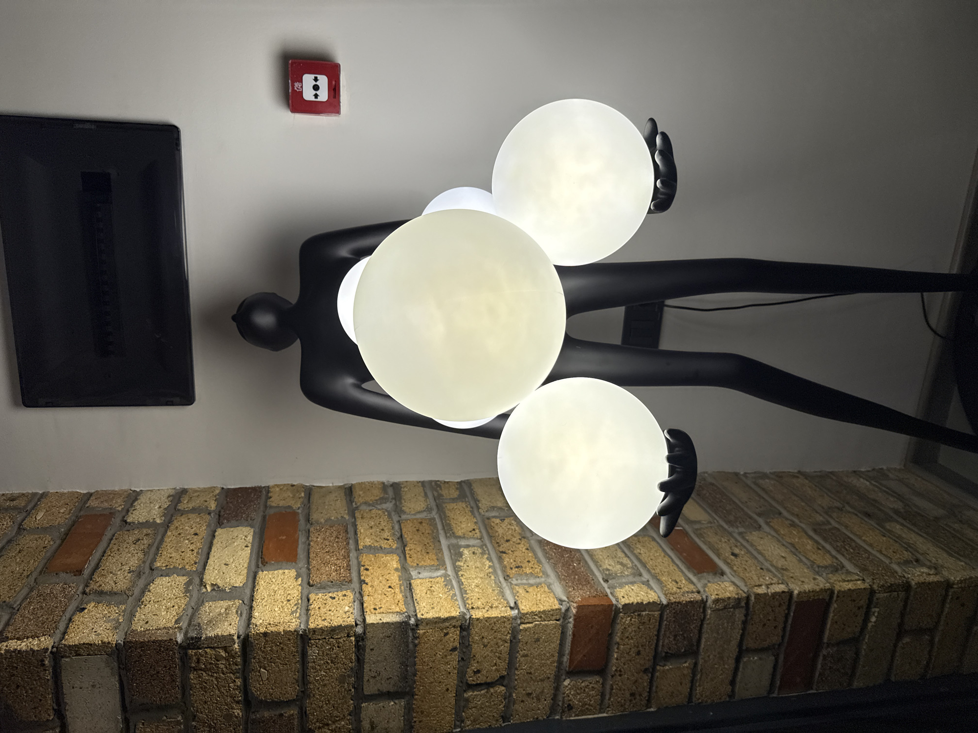 An interior scene with a brick column on the left. A black, human-shaped floor lamp with multiple glowing white spheres held in its hands is positioned in the center. On the wall behind the lamp, there is a black electrical panel at the top and a red fire alarm box on the right. The background wall is painted a light color, and the floor is dark. The lighting is dim, with the spheres on the lamp being the primary light source. The location is stated to be in Dilijan, Armenia.