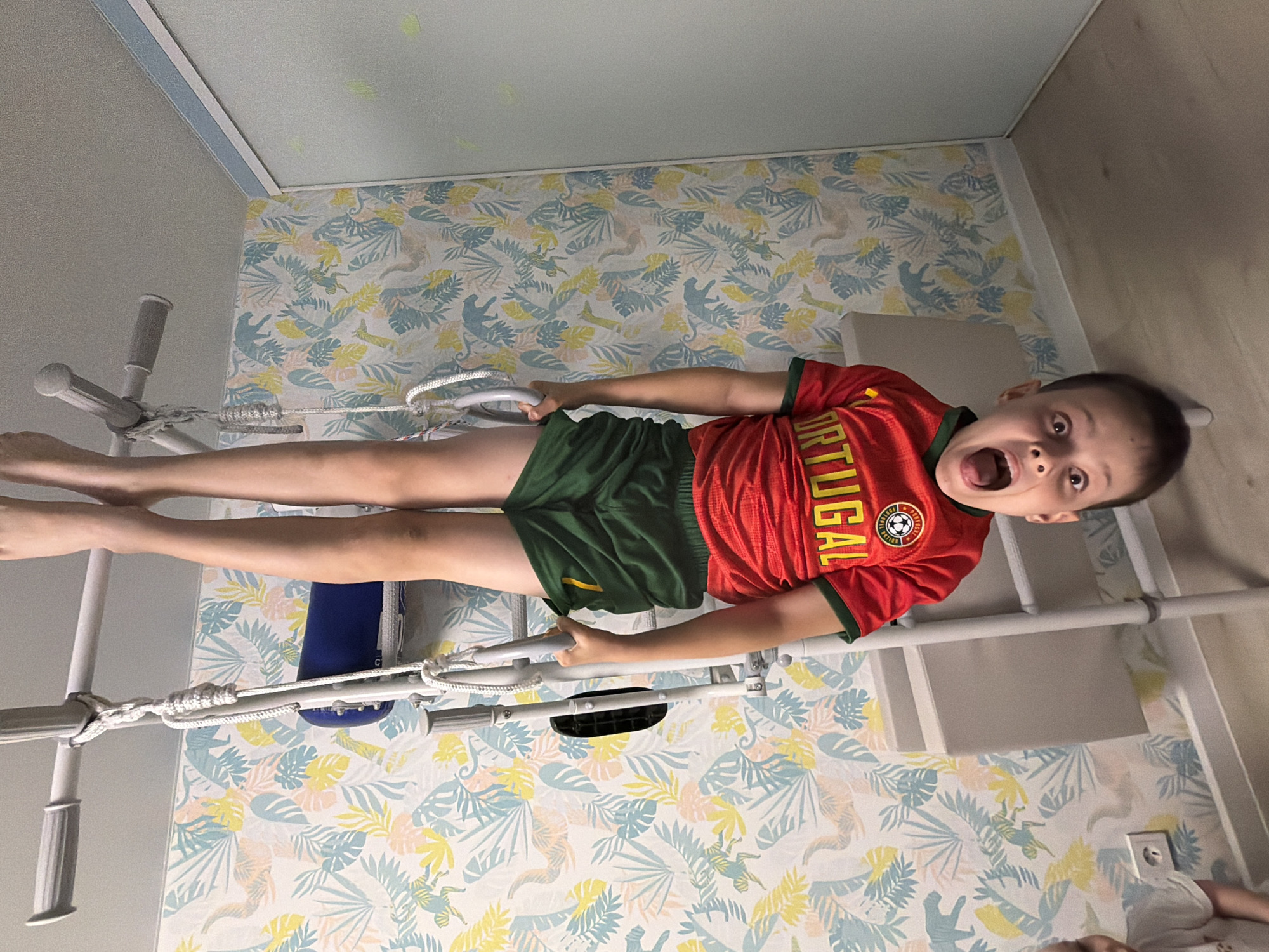 The image, rotated 90 degrees clockwise, captures a young boy horizontally suspended on a white indoor climbing frame within a room in Votkinsk, Russia. He is actively holding onto the rungs with both hands, his legs extended, suggesting an exercise or playful stunt. The boy is wearing a red football (soccer) jersey with "PORTUGAL" emblazoned vertically on the side, accompanied by a small crest featuring a football design, and green athletic shorts. His face is expressive, with wide-open eyes and a gaping mouth, conveying a vivid sense of surprise, excitement, or playful exertion.
The background wall is adorned with a vibrant wallpaper featuring a light blue, yellow, and beige tropical or jungle pattern with stylized dinosaurs and foliage. The ceiling above has a light blue trim. To the right, a section of a light wooden-paneled wall is visible. Elements of the climbing frame, including ropes and various rungs, are prominent. In the lower right corner of the original image, a white electrical outlet is visible, and part of another individual, possibly a child, lying down, can be faintly seen. The scene is brightly lit, indicative of an indoor setting during daytime.