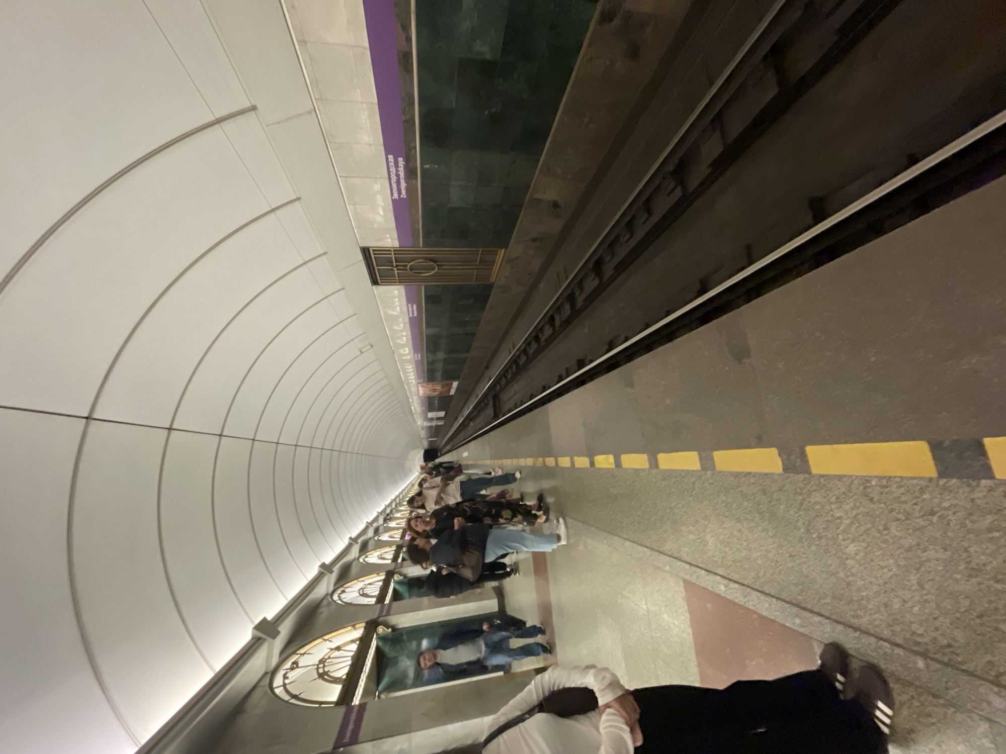 The image shows the interior of a Saint Petersburg Metro station, specifically Zvenigorodskaya station, as indicated by the bilingual signage on the wall. The station features a long, curved tunnel with a vaulted ceiling. The platform is lined with a granite-like material and marked with yellow safety lines. On the platform, several individuals are standing and waiting, some are looking at their phones. To the right, train tracks are visible. The station's architecture includes arched openings with decorative elements and a wall clad in dark green marble-like tiles. The overall atmosphere is typical of an underground metro station.