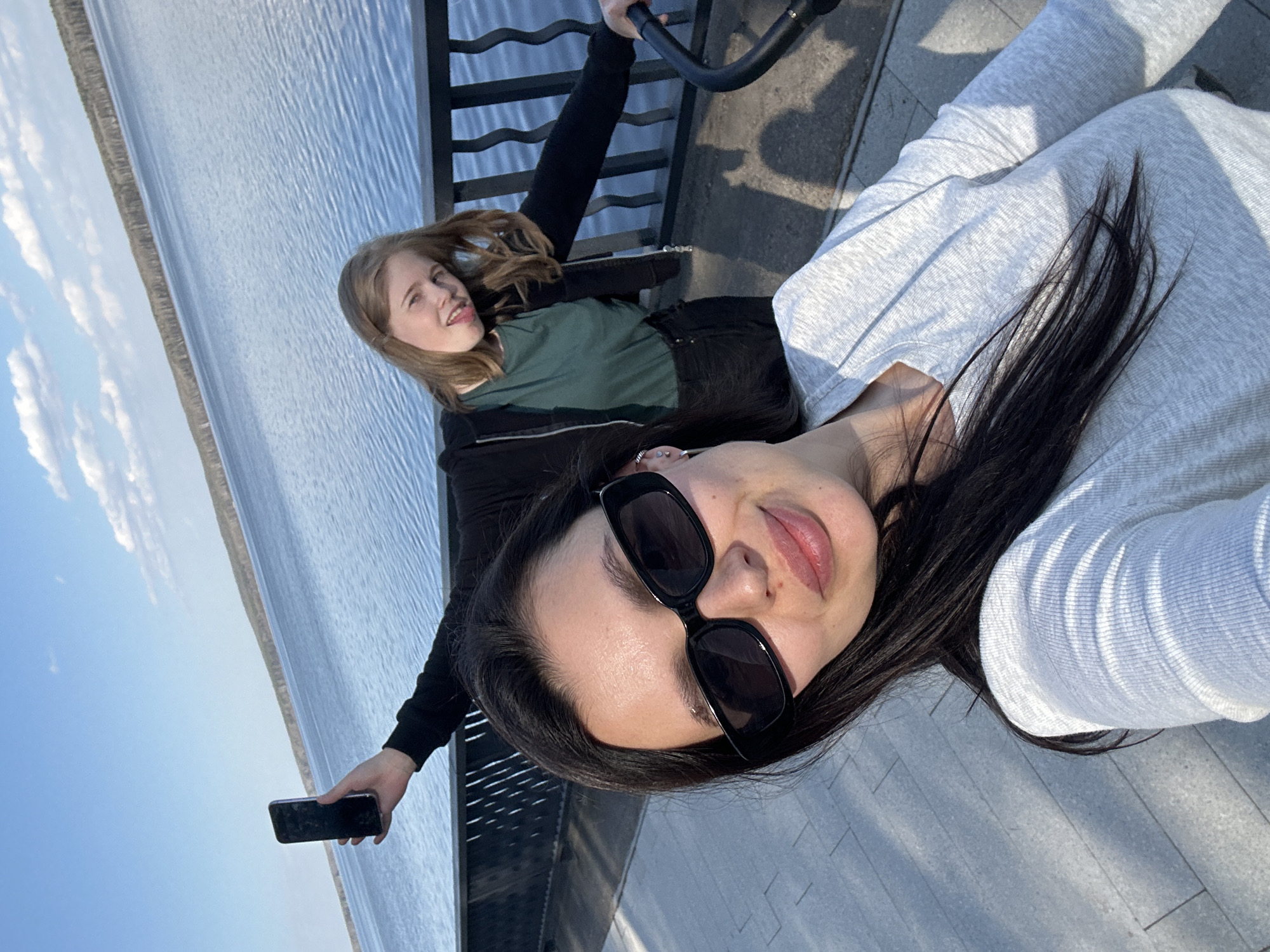 A photograph, taken at a tilted angle, depicts two individuals, presumably at a waterfront location in Chaikovsky, Russia, given the context. The person closest to the camera is a woman wearing sunglasses and a light grey long-sleeved shirt. Her dark hair is visible. Behind her, a younger person with blonde hair wears a green shirt and black jacket. The younger person is near a railing. The woman holds a phone in her left hand. The background shows a body of water, and the sky, suggesting an outdoor setting.