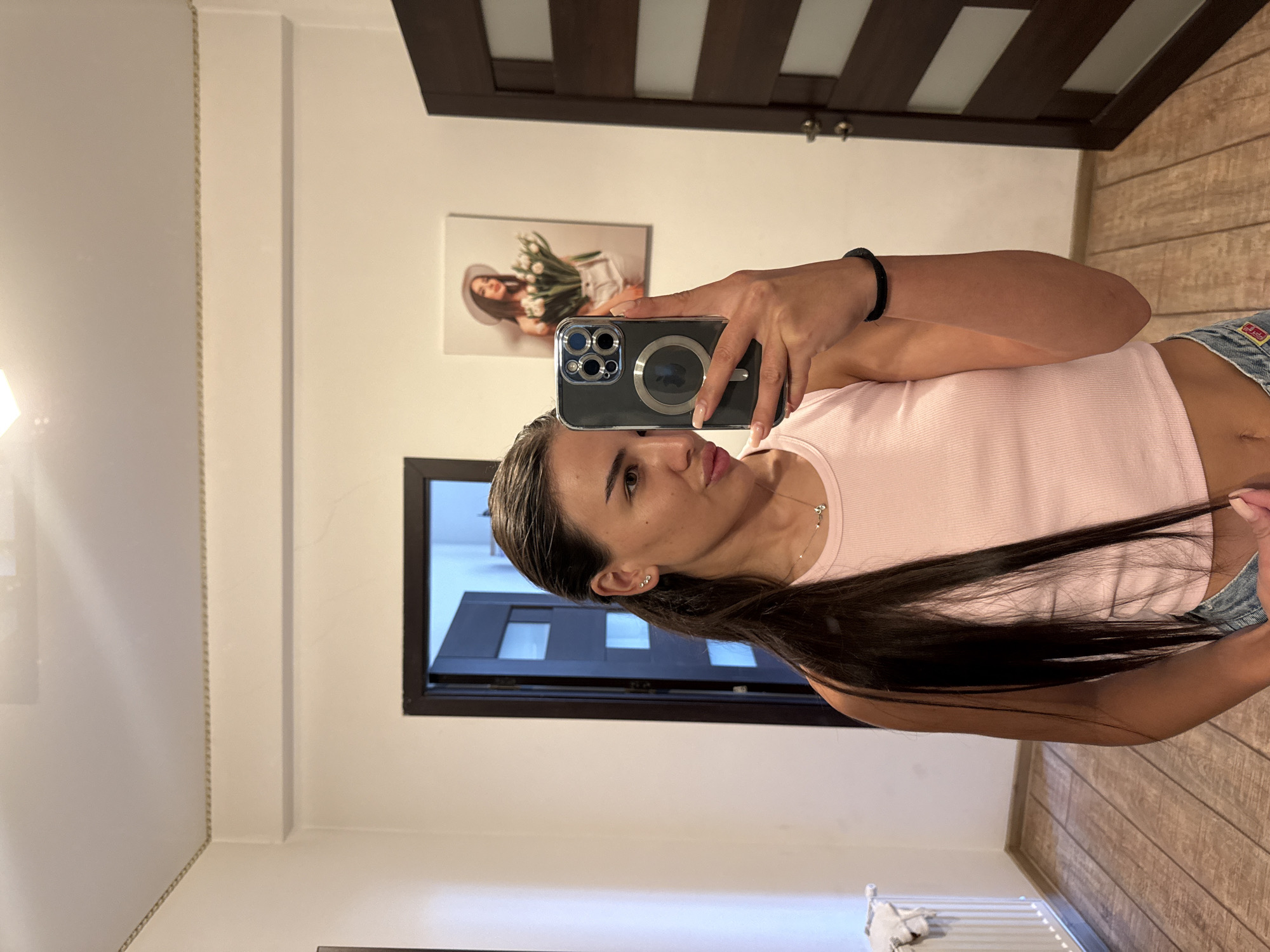 The image shows a young woman taking a selfie in a room that appears to be a hallway or a dressing room. She is wearing a light pink ribbed crop top and denim shorts. Her long, dark hair is pulled back into a ponytail. She is holding a smartphone in her right hand, which is angled to capture her reflection. In the background, there is a framed picture of a woman wearing a hat and holding a bouquet of flowers. There is also a door on the right side of the image with horizontal glass panels. On the left side, a radiator is visible. The flooring is made of wooden planks. The woman is wearing simple earrings and a delicate necklace. The overall setting suggests a domestic indoor environment. There is no information within the image to suggest the location is in Chișinău, Moldova.