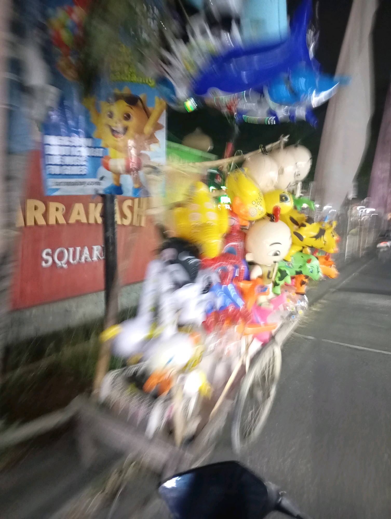The image depicts a blurry nighttime scene in Babelan, Indonesia. A cart filled with colorful balloons is prominently displayed, featuring various character-shaped balloons such as a shark, a baby, and animal figures. The cart is positioned in front of a red sign with the words "ARRAKASH SQUARE" written in white capital letters. The scene appears to be outdoors, with a dark background suggesting it is nighttime. The motion blur indicates movement, likely of the cart or the person taking the photograph.