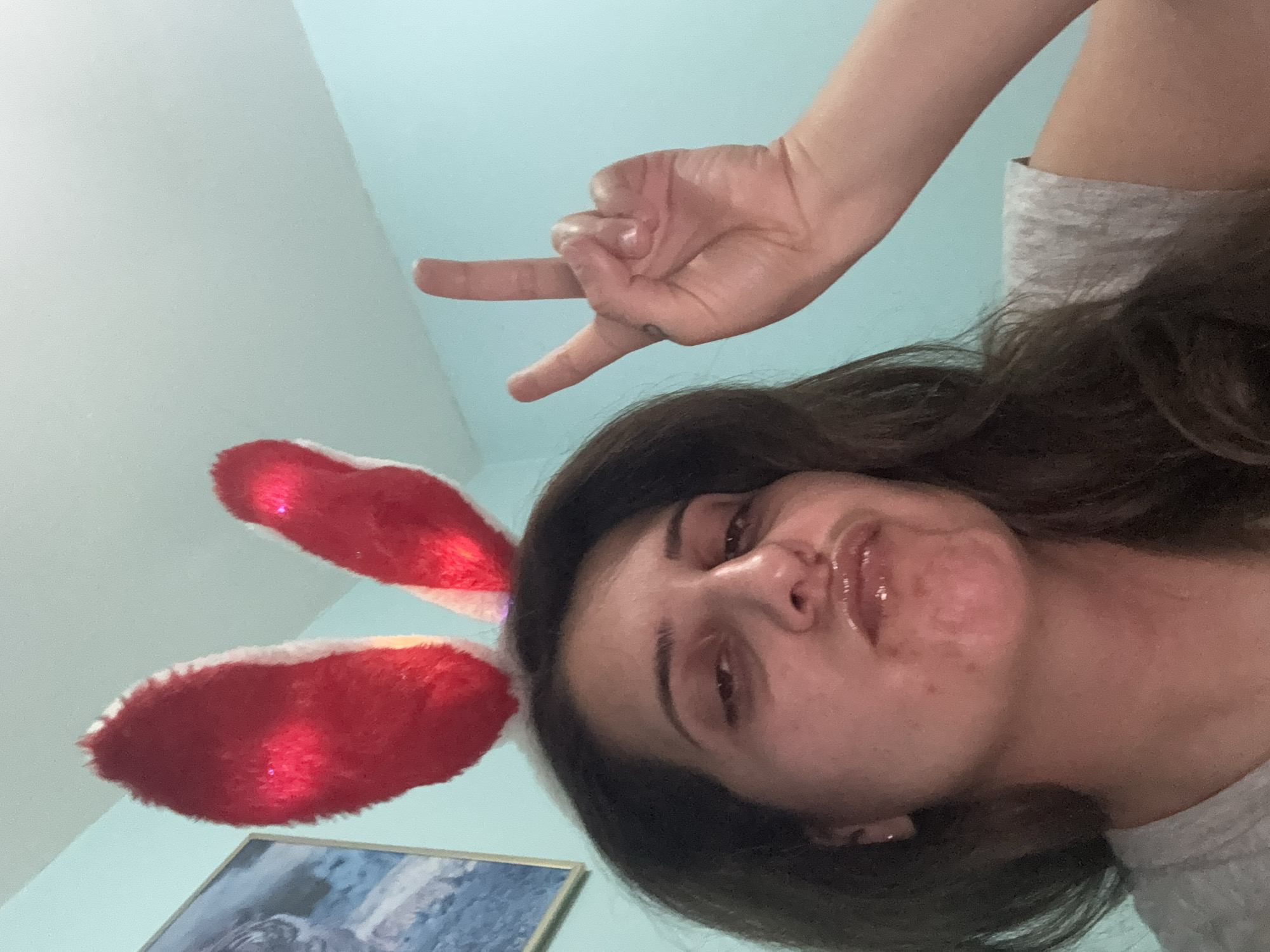 A woman is taking a selfie, wearing red bunny ears with lights and making a peace sign with her right hand. She is wearing a grey t-shirt. The background is a pale blue wall. There is a framed picture on the wall to the left of the woman. This image was taken indoors.