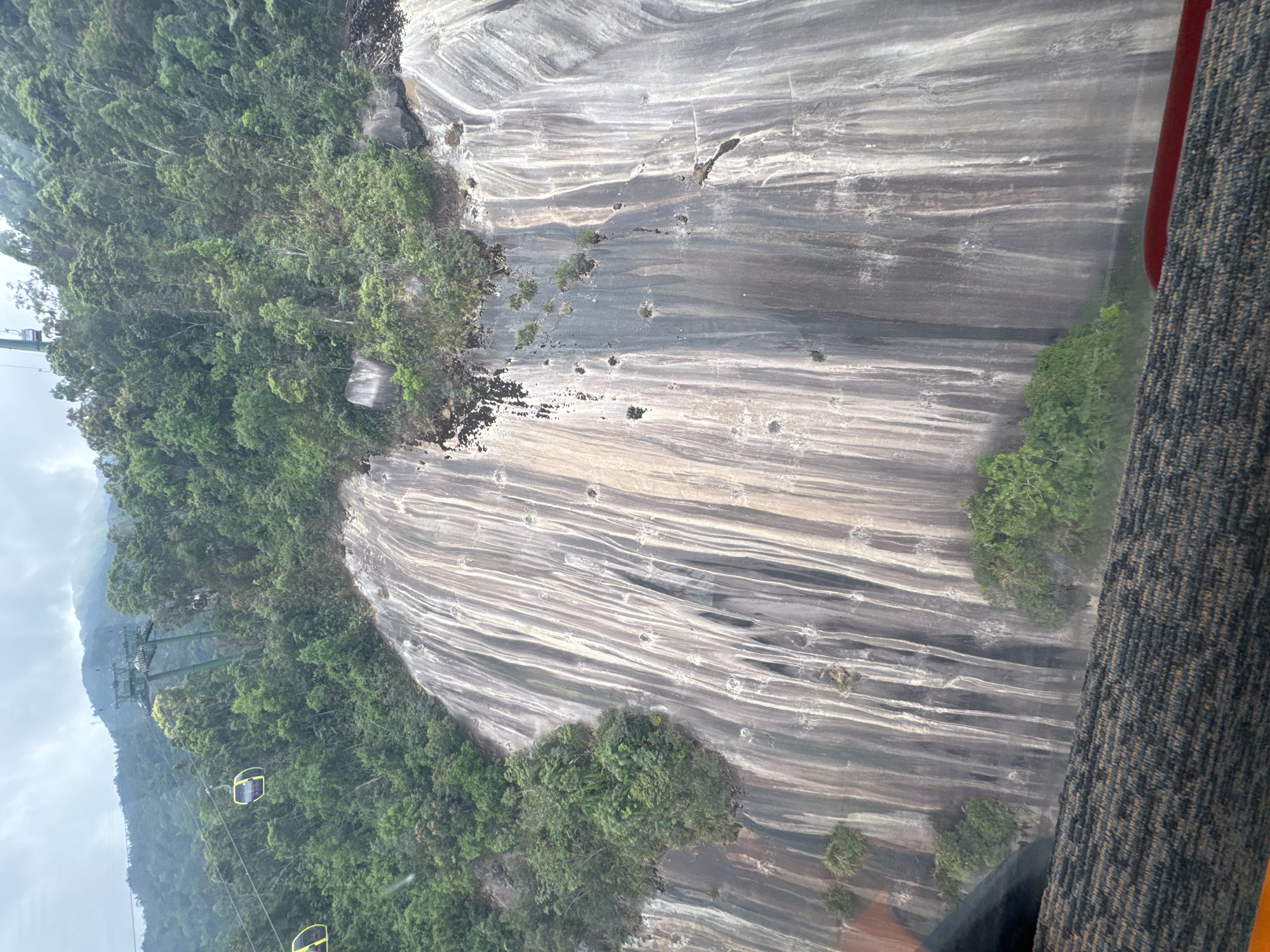 View from inside a cable car ascending Ba Na Hills in Đà Nẵng, Vietnam. The cable car system is visible, with several cars traversing the misty landscape. Dense green vegetation clings to the side of a steep, striated rock face. The foreground includes a portion of the cable car interior, featuring a patterned textile.