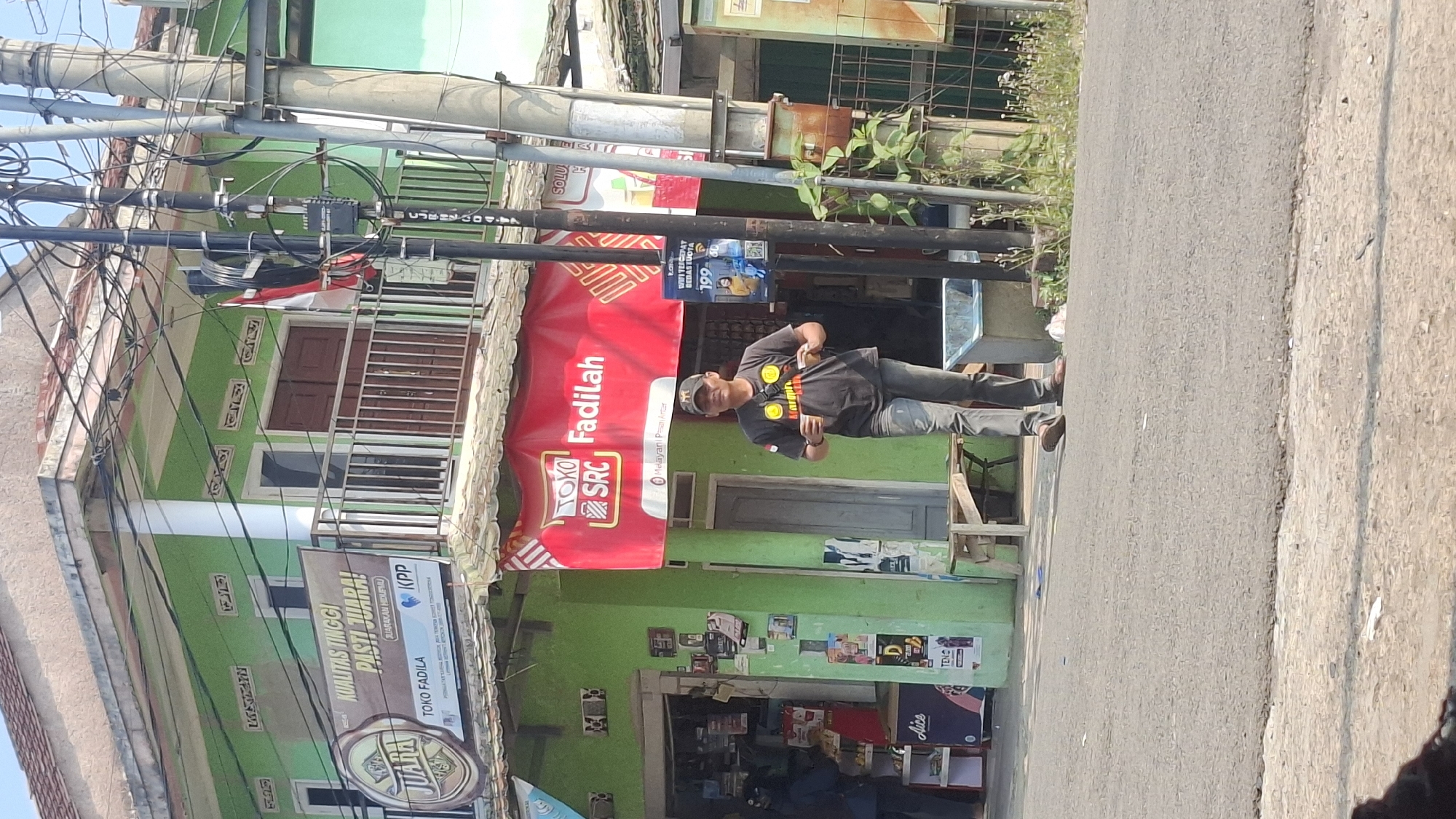 The image depicts a street scene in Cilaku, Indonesia, likely on a sunny day given the shadows. In the foreground, a man wearing a dark t-shirt with yellow graphics and a baseball cap is walking on the paved road, holding a cup in each hand. He is wearing gray pants and sandals. Behind him, a green building with a red banner that reads "TOKO SRC Fadilah" is visible. The banner also displays "Melayani Pesan Antar." The building appears to be a store or small business. To the left of the building, a smaller shop with shelves stocked with products is partially visible. A wooden bench is placed outside the green building. In the background, utility poles with numerous wires are prominent, and a flag, possibly the Indonesian flag, is attached to one of them. The environment suggests a semi-urban or rural setting with modest architecture.