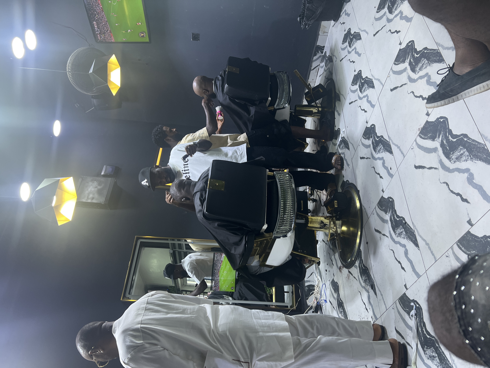 The image captures a scene inside a barbershop located in Lugbe, Nigeria. Several individuals are present, including barbers and customers. Two barbers are actively attending to customers seated in barber chairs. A third man, dressed in a white kaftan, stands in the foreground. Overhead lighting fixtures and a TV screen showing a soccer game are visible. The floor is tiled with a black and white wave-like pattern.