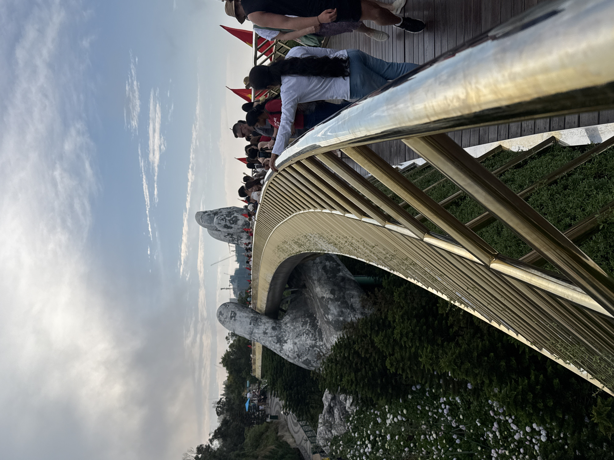 The image presents a vibrant scene on the Golden Bridge (Cầu Vàng) at Ba Na Hills in Da Nang, Vietnam, an iconic pedestrian bridge known for its unique design. The bridge features a shiny, golden-colored railing and a wooden pathway, supported dramatically by colossal, weathered grey stone hands that appear to cradle it above the lush landscape.
Numerous people, primarily tourists, are present on the bridge. They are seen walking, standing, and leaning on the railing, observing the panoramic views. In the foreground, a woman with long dark hair, wearing a white shirt and blue jeans, leans on the golden railing, looking out. Other visitors are visible further along the bridge, some engaging in sightseeing or taking photographs.
The setting is an elevated outdoor environment, likely mountainous, with dense green foliage and small white flowers bordering the bridge structure. The sky is bright with patches of blue and scattered white, wispy clouds, suggesting fair weather during what appears to be the late afternoon or early evening, given the soft light that reflects off the golden surfaces. Red flags adorned with yellow stars, representing the national flag of Vietnam, are interspersed along the bridge, providing a clear location cue. The overall atmosphere is one of active sightseeing and enjoyment at a popular tourist destination. No legible text is visible on the bridge itself or any other objects, apart from the symbolic elements on the flags.