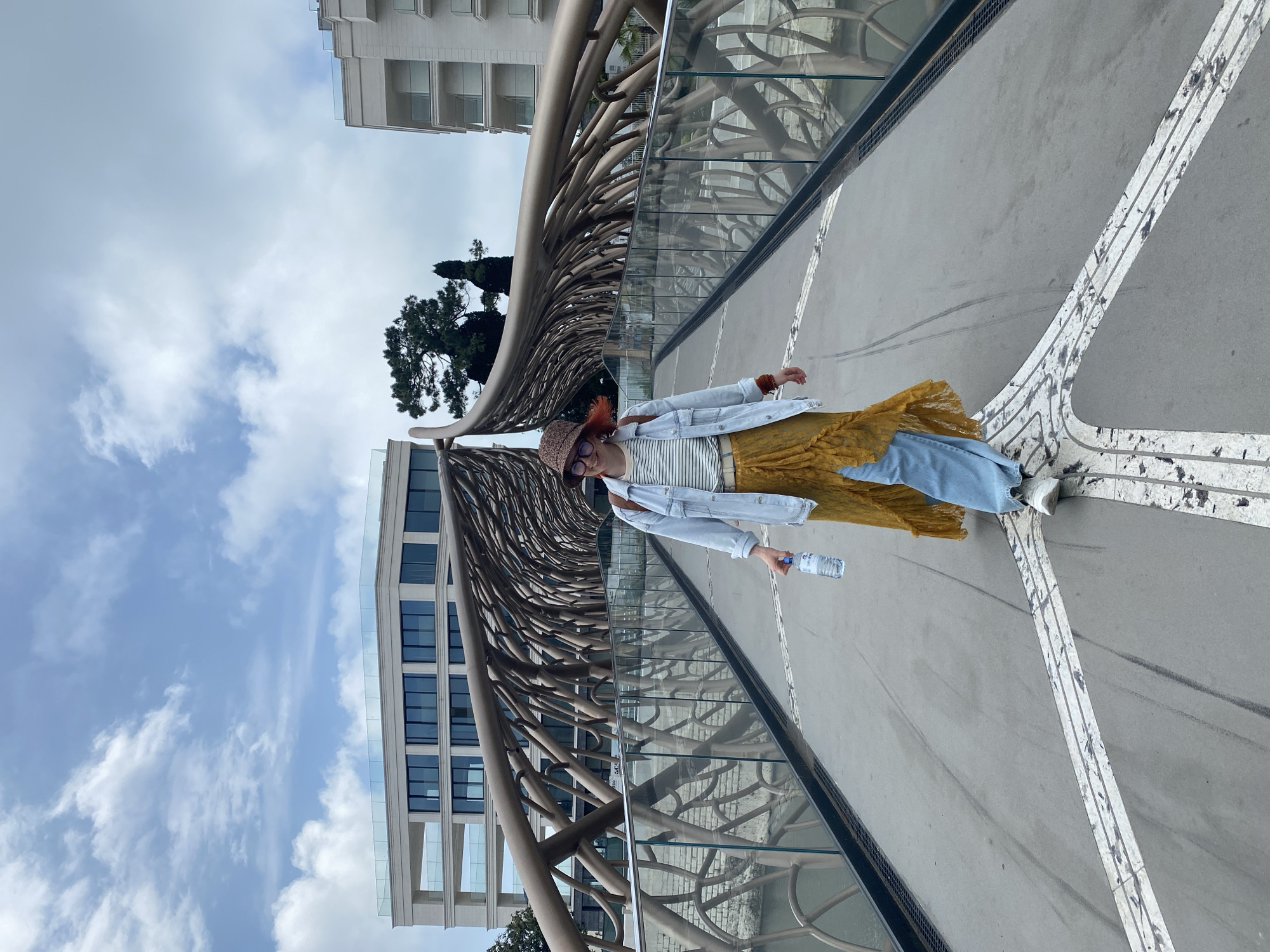 A woman with red hair and a straw hat stands on a bridge in Sochi, Russia. She wears a striped shirt, a yellow skirt, and a light blue denim jacket. She holds a bottle of water in her right hand. The bridge features an intricate metal structure that resembles tree branches. Glass panels line the sides of the bridge. A white building with multiple windows is visible in the background, along with a blue sky and clouds. The bridge surface is gray with white painted markings.