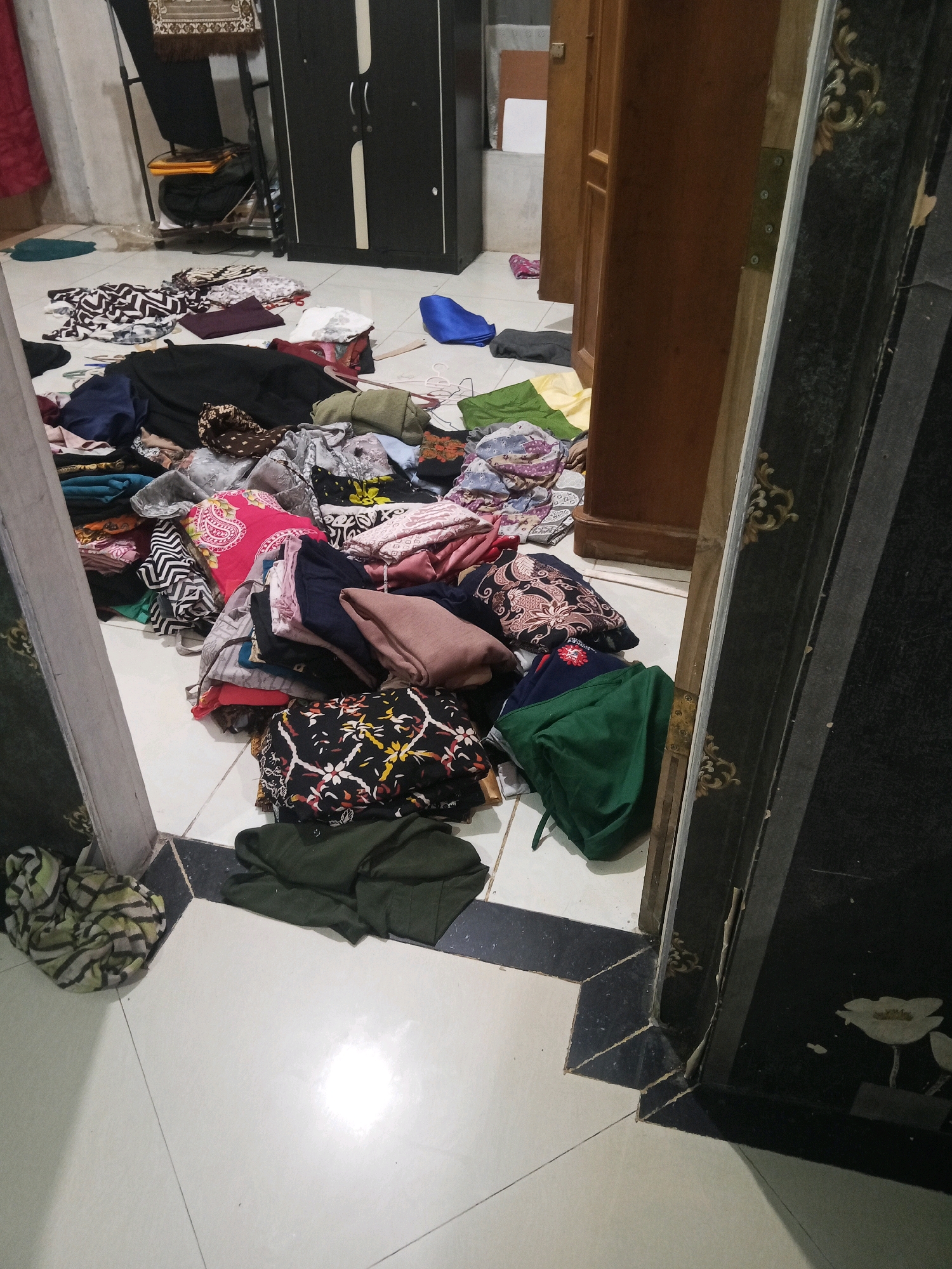 A cluttered room floor in what appears to be a residential dwelling in Jinang Kampung Pansur, Indonesia, is covered with a large pile of folded and unfolded clothing. The clothing consists of various garments in different colors and patterns, including shirts, pants, and possibly dresses. Some items are neatly folded, while others are scattered loosely. The floor is tiled with a combination of white and black tiles, forming a geometric pattern. In the background, a dark wardrobe with double doors is visible, as is a wooden doorframe and a portion of a dark-colored decorative panel with floral motifs. The lighting in the room is dim, suggesting it might be evening or the curtains are drawn. The overall scene conveys a sense of disarray and a lack of organization, possibly indicating laundry that has not yet been put away or is in the process of being sorted. There are no people or animals visible in the image.