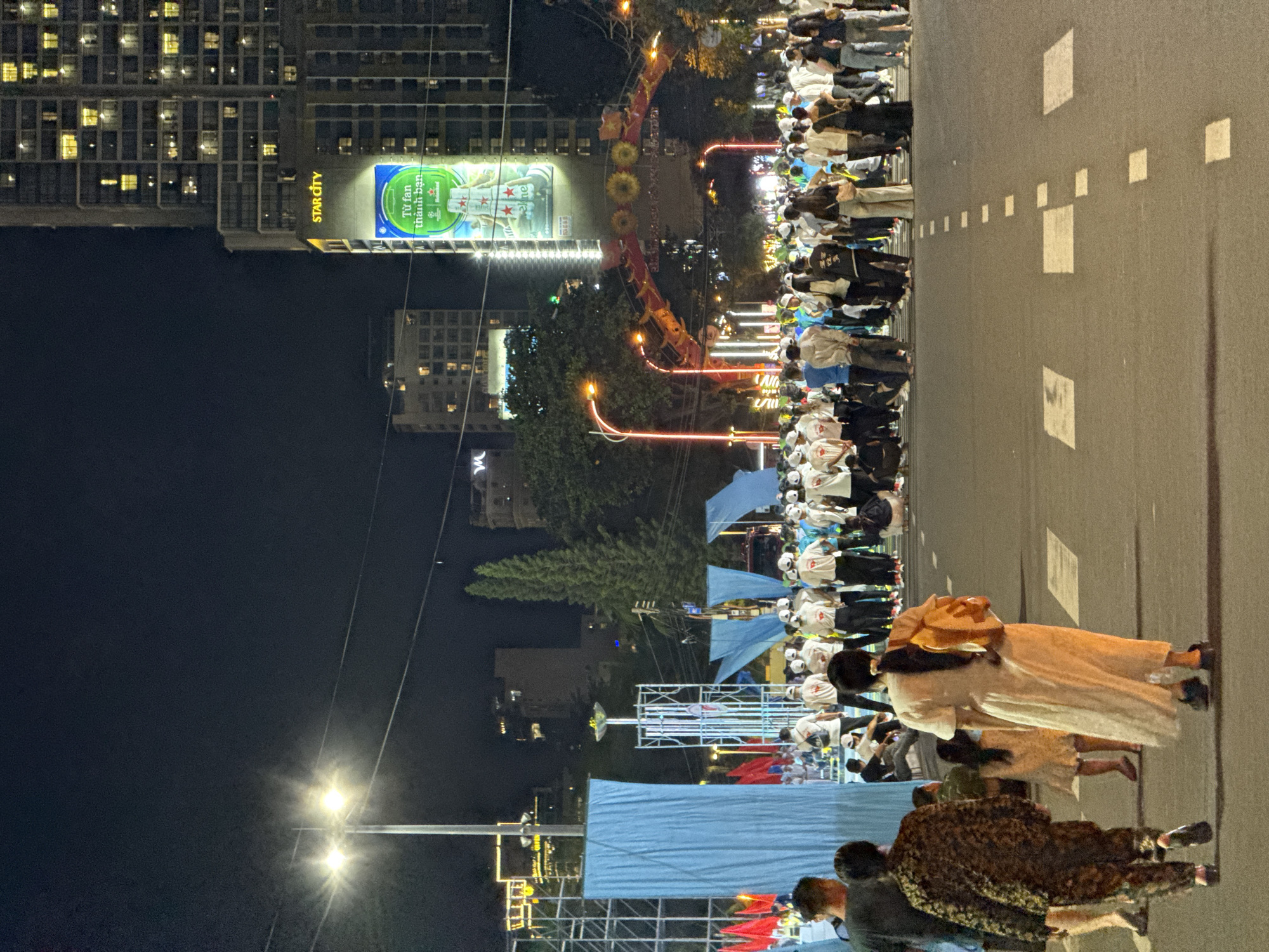 A nighttime street scene in Nha Trang, Vietnam, captures a large gathering of people walking and standing on a wide, paved road. The scene is illuminated by streetlights and the glowing lights of a tall, modern building on the right, identified by a "STAR CITY" sign and a large billboard displaying a Heineken advertisement with Vietnamese text. In the background, an illuminated archway with decorative elements suggests a festive or event-related atmosphere.
The foreground features several individuals walking away from the camera. A woman in a long, white dress and an orange backpack is prominent, followed by a child in a patterned dress. To their left, another individual in a camouflage-patterned outfit walks ahead. A crowd of people, many wearing white t-shirts and hats, are gathered further down the street, some appearing to be waiting or participating in an organized event. The road itself is marked with white squares and dashed lines, indicating a pedestrian crossing or traffic lane. The overall environment suggests an urban setting at night, likely a public space or thoroughfare within the city of Nha Trang.