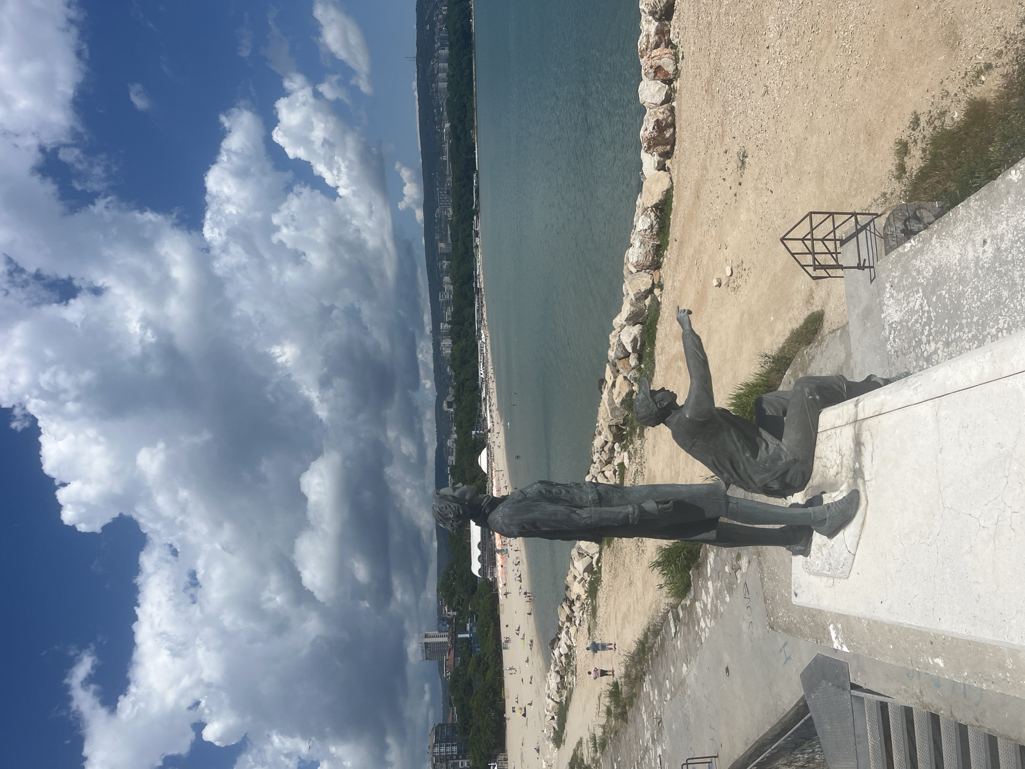 This image depicts a view of the Black Sea coast in Varna, Bulgaria. In the foreground, two bronze statues stand on a concrete pedestal. One statue is of a standing figure, facing away from the viewer and looking towards the sea. The other statue is of a seated figure, pointing out towards the sea and horizon.
Beyond the statues, a sandy beach stretches along the coastline, with scattered beachgoers visible. The water is a clear blue-green, and in the distance, the cityscape of Varna rises, with buildings and greenery lining the hills. The sky is a bright blue with dramatic, fluffy white clouds.
The location is a prominent spot along the Varna coastline, likely part of the Sea Garden or a similar public area overlooking the beach. The statues suggest a historical or commemorative aspect to the site. The overall scene captures a sunny day at the beach, with a blend of natural scenery and urban development.