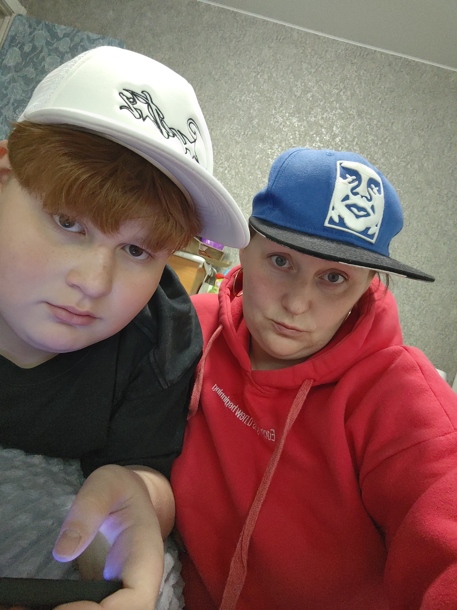 The media file captures two individuals posing for a close-up photograph indoors, likely in a casual home setting in Markovskii, Russia.
On the left is a person with reddish-brown hair, wearing a white trucker hat with a stylized black design on the front, and a black top or hoodie. They are holding a device, possibly a smartphone, which casts a blue light onto their hand. This individual has a neutral expression, looking directly at the camera.
To their right is an adult female wearing a red hooded sweatshirt with a drawstring and a blue baseball cap featuring a white "Obey Giant" logo (a stylized face). She has gold hoop earrings and a slightly serious expression with pursed lips. The text on her red hoodie, visible in reverse due to the image orientation, reads "Emergency World".
The background consists of a light grey wall with a subtle mottled pattern. Both subjects are looking towards the camera, suggesting they are posing for a selfie or a directed photograph in an informal, relaxed environment.
