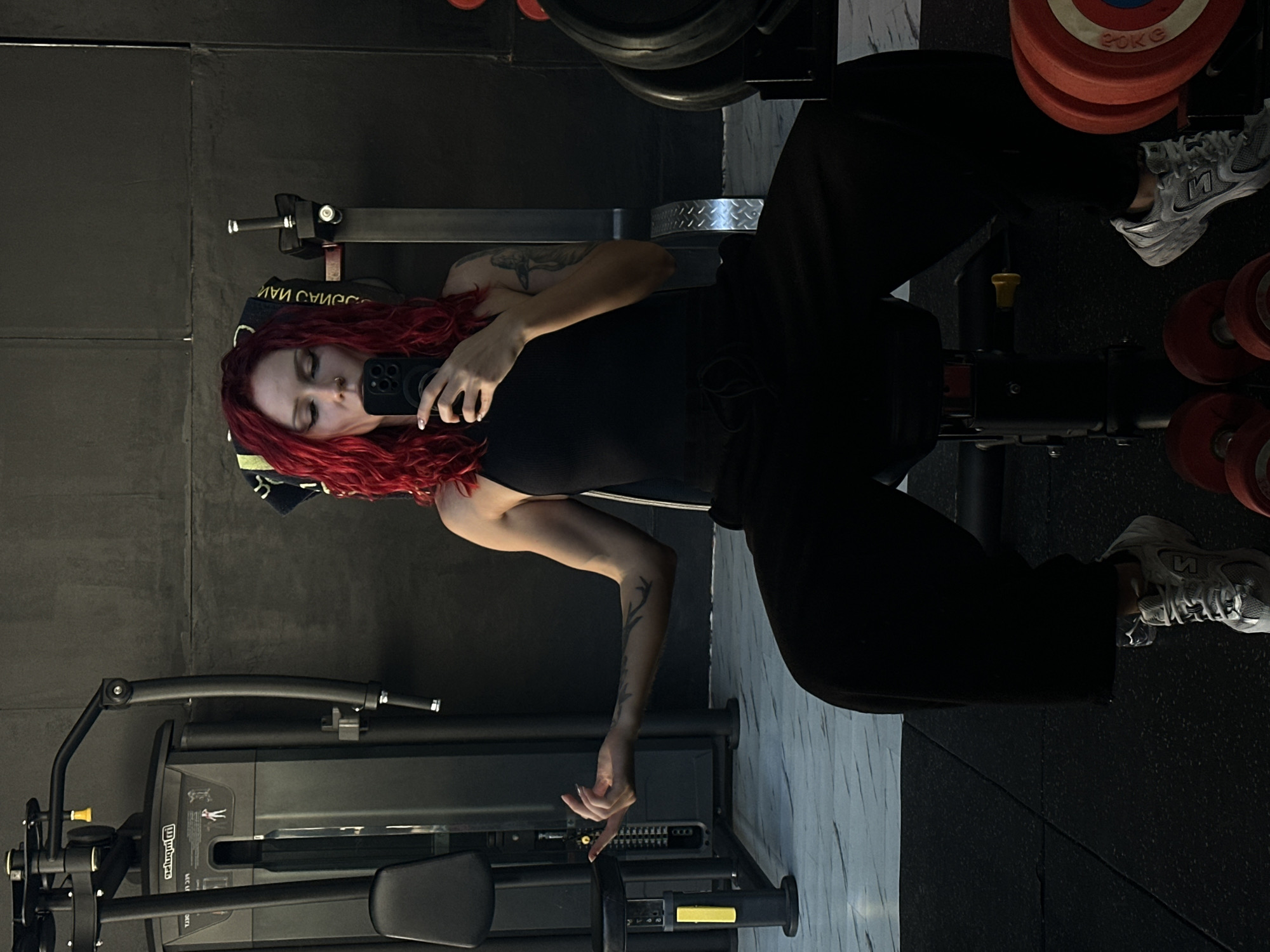 A woman with red hair is in a gym, lying on a weight bench, holding a black cell phone. She is wearing a black tank top and baggy black pants, with grey New Balance sneakers. The gym has black walls, marble-patterned flooring, and weight machines are visible, along with red weights. The location is possibly North Kuta, Indonesia.