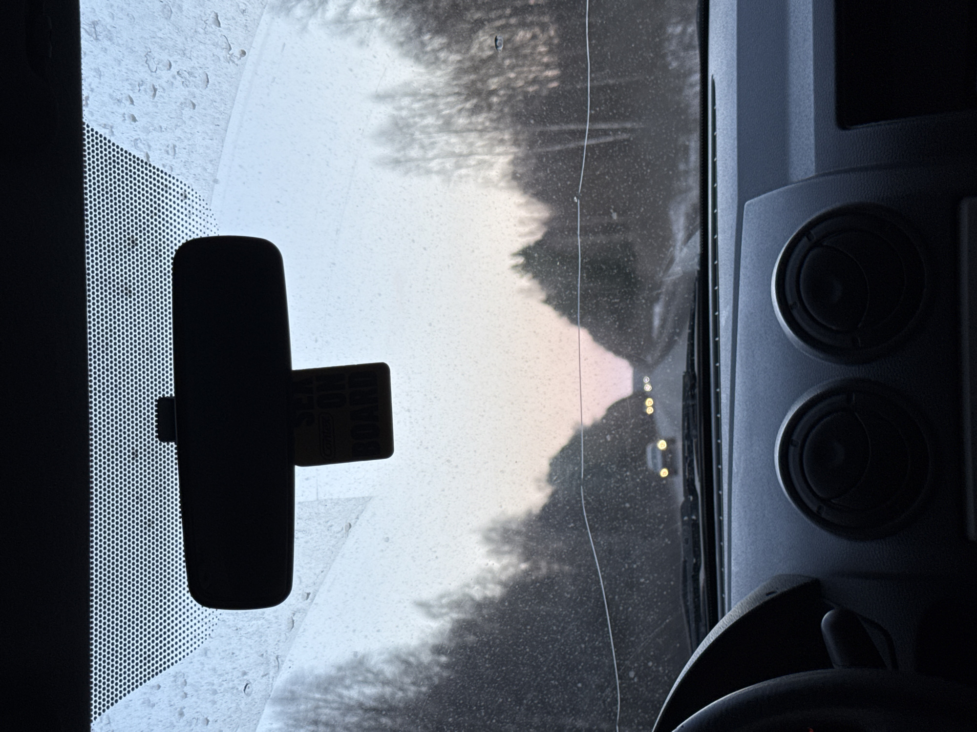 The image is a view from inside a vehicle, likely a car or truck, looking out through the windshield. The interior of the vehicle, including the dashboard, steering wheel, and rearview mirror, are visible. The windshield has a crack running down the center. Outside the vehicle, trees line what appears to be a road, with the hazy light suggesting either dawn or dusk. There are lights visible in the distance, indicating other vehicles. There is no verifiable information to suggest a specific location.