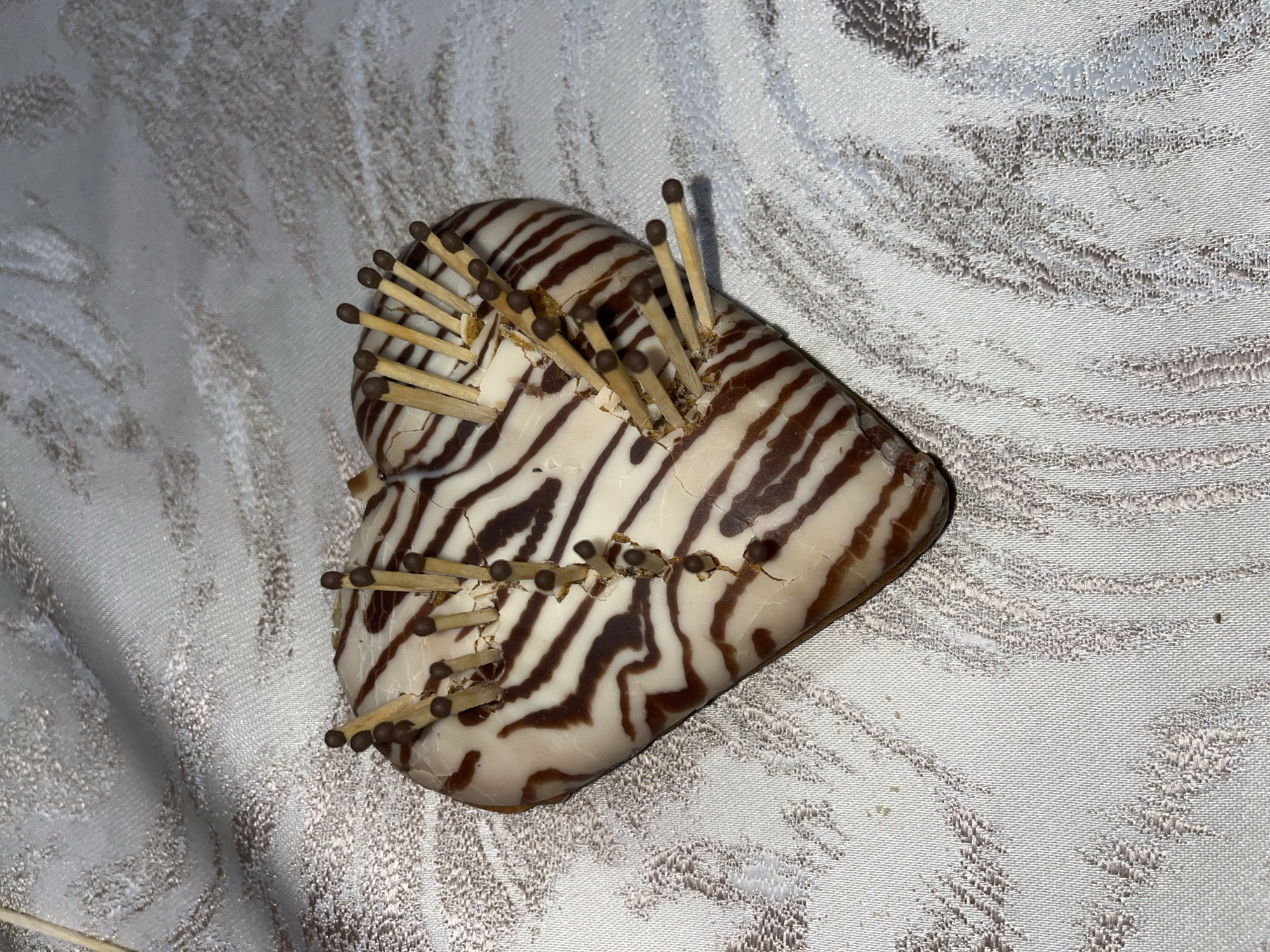 A heart-shaped object with brown and white zebra-like stripes is positioned on a light-colored fabric with a patterned texture. Multiple wooden matchsticks are stuck into the object, resembling needles in a pin cushion. The background fabric exhibits a swirled pattern. The location is Lozarevo, Bulgaria.