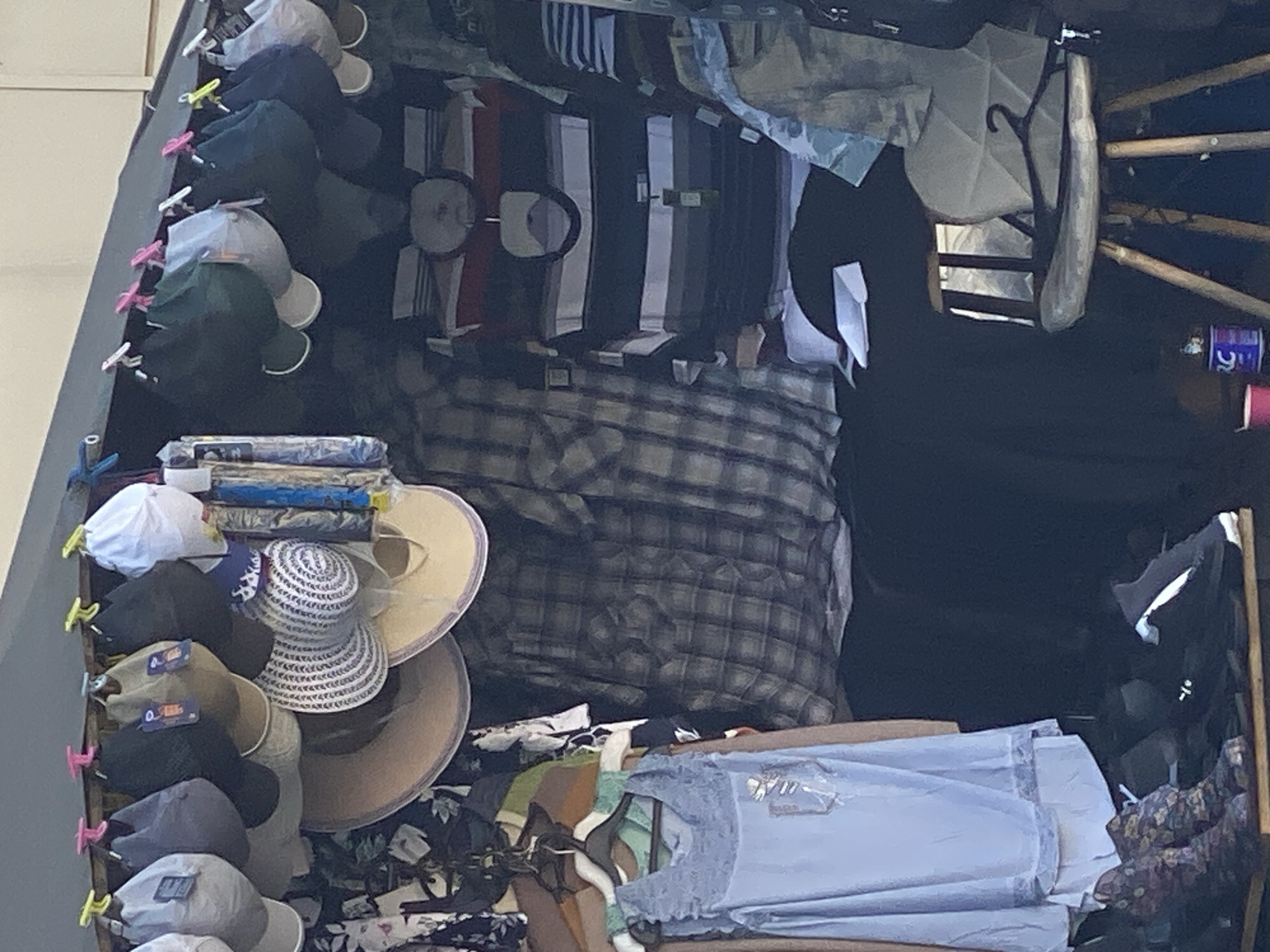 This is a forensic, matter-of-fact description of the provided image.
The image depicts a stall selling clothing and hats, likely in an outdoor market. The stall is covered by a dark tarp or awning. Hanging from a rail at the top are numerous hats of various styles and colors, including baseball caps and wider-brimmed hats. Below the hats, various articles of clothing are displayed on hangers and folded. Among these are at least two plaid shirts, one light blue sleeveless top with ruffles, and a horizontally striped long-sleeved shirt. There are also what appear to be bags or folded fabric items further back. On a lower shelf or display, a pair of patterned boots and a black sneaker are visible. A wooden stool with a cushioned seat and visible legs is situated to the right of the clothing display. Two containers, one pink and one blue with white lettering, are on the ground near the stool. The overall lighting suggests it is daytime. There are no visible people in the image, and no specific identifying features indicate the location is Novokuznetsk, Russia.