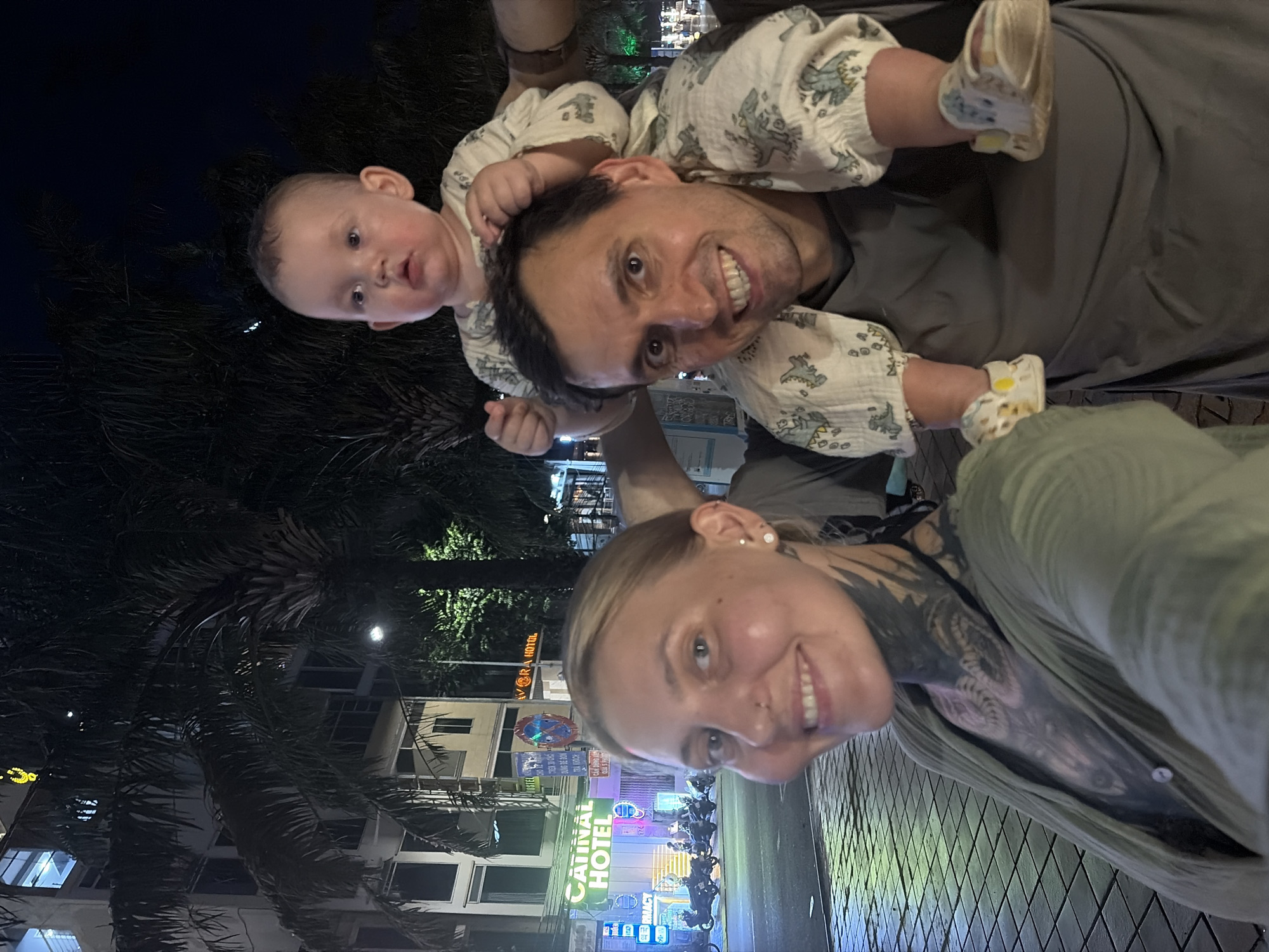 This is a nighttime selfie-style photograph capturing a family of three – a man, a woman, and a baby – in an urban outdoor setting in Đà Nẵng, Vietnam.
The woman, with light hair and visible tattoos on her left arm/shoulder, is in the foreground, holding the camera and smiling broadly. Next to her, a man carries a baby on his shoulders, also smiling towards the camera. The baby, dressed in a light-colored outfit with dinosaur patterns and small white and yellow shoes, looks directly at the camera with a curious expression.
The background features dark palm trees and several illuminated buildings, indicating an evening scene. Bright neon signs are visible, including "CATALINA HOTEL" in green letters, "VORA HOTEL," and a partially visible "PHARMACY" sign. Other unreadable Vietnamese text can be seen on street signs and advertisements. Several motorcycles are parked on the street in the middle ground, suggesting a bustling location. The overall atmosphere conveyed by the smiling faces is one of happiness and enjoyment during an outing.