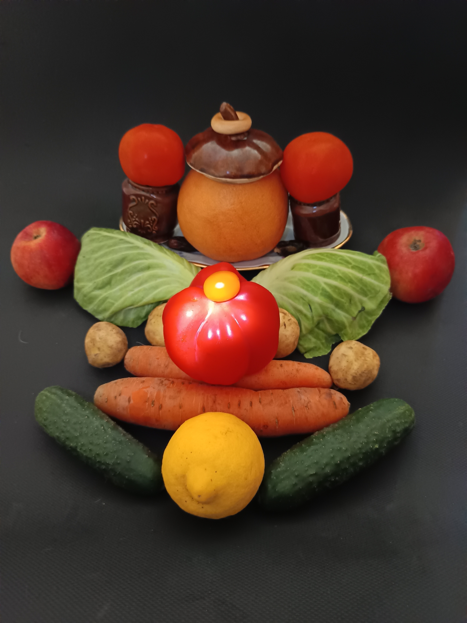 A still life arrangement of vegetables and ceramic pots is displayed against a dark, textured background. The composition is symmetrical, with a central, round, bright red bell pepper topped with a small, illuminated yellow object, possibly a candle or light. Flanking the bell pepper are two whole cabbages. Below the cabbages, arranged in a curved pattern, are four potatoes and three carrots. At the bottom of the arrangement, two cucumbers and a bright yellow lemon are positioned in a semi-circle.
In the background, a round, orange-toned ceramic pot with a brown lid sits centrally. To its left and right are smaller, darker brown ceramic pots or jars, each topped with a bright red tomato. The ceramic items rest on a white plate with a gold rim. The lighting appears to be from above and slightly to the front, creating highlights on the vegetables. The dark background suggests an indoor setting, possibly a table or surface in a room with controlled lighting. The overall impression is one of abundance and rustic charm. No text is visible in the image.