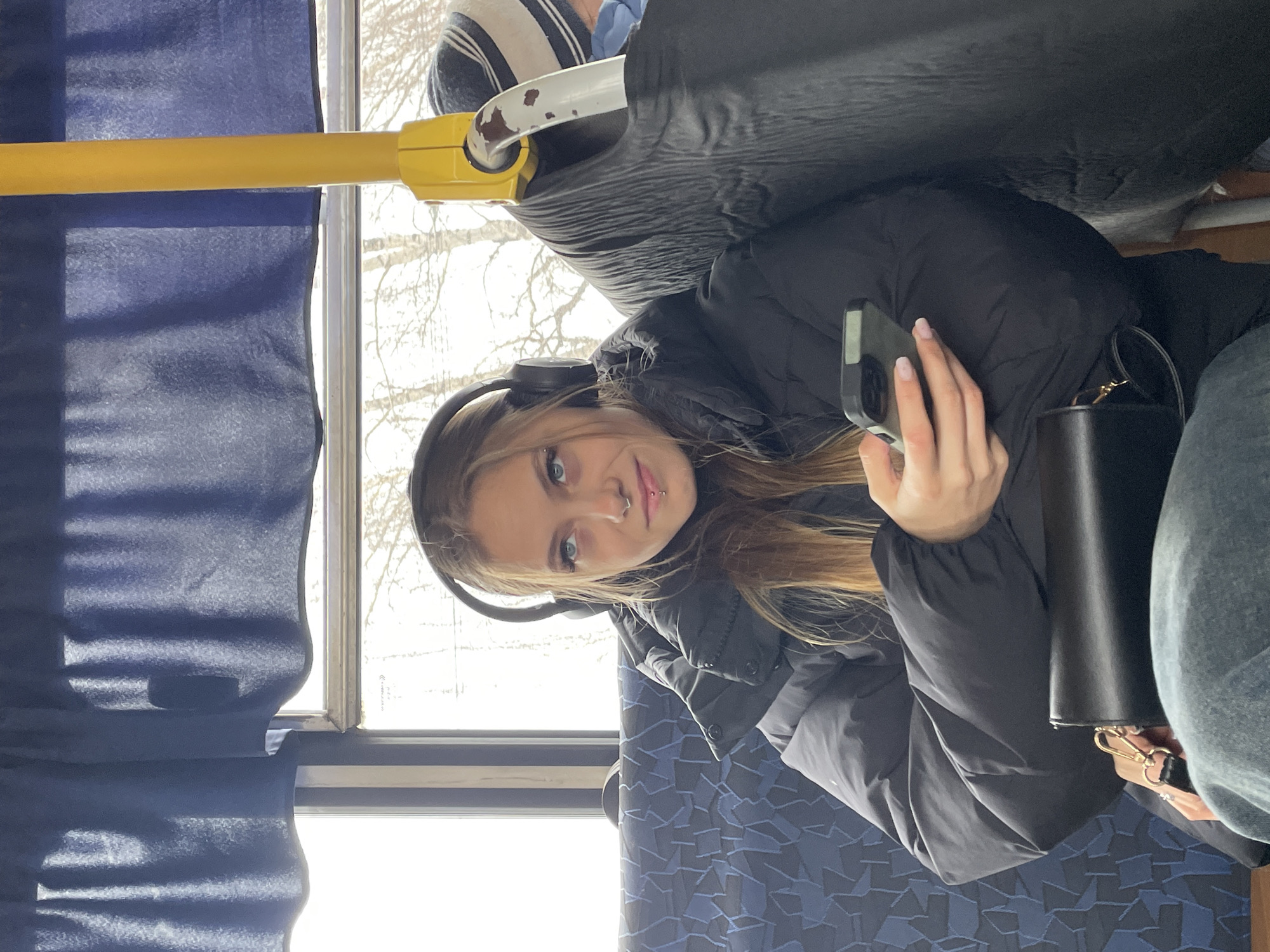 A young woman with light brown hair and blue eyes, wearing headphones and a black puffer jacket, is seated on public transport, possibly a bus, holding a smartphone in her right hand. She has a nose piercing and a lip piercing. Next to her is a black handbag and a person wearing gray pants. A yellow handrail is visible above, and a window shows a view of trees. The interior of the bus has blue patterned seats and blue curtains. The location is Фоки, Russia.