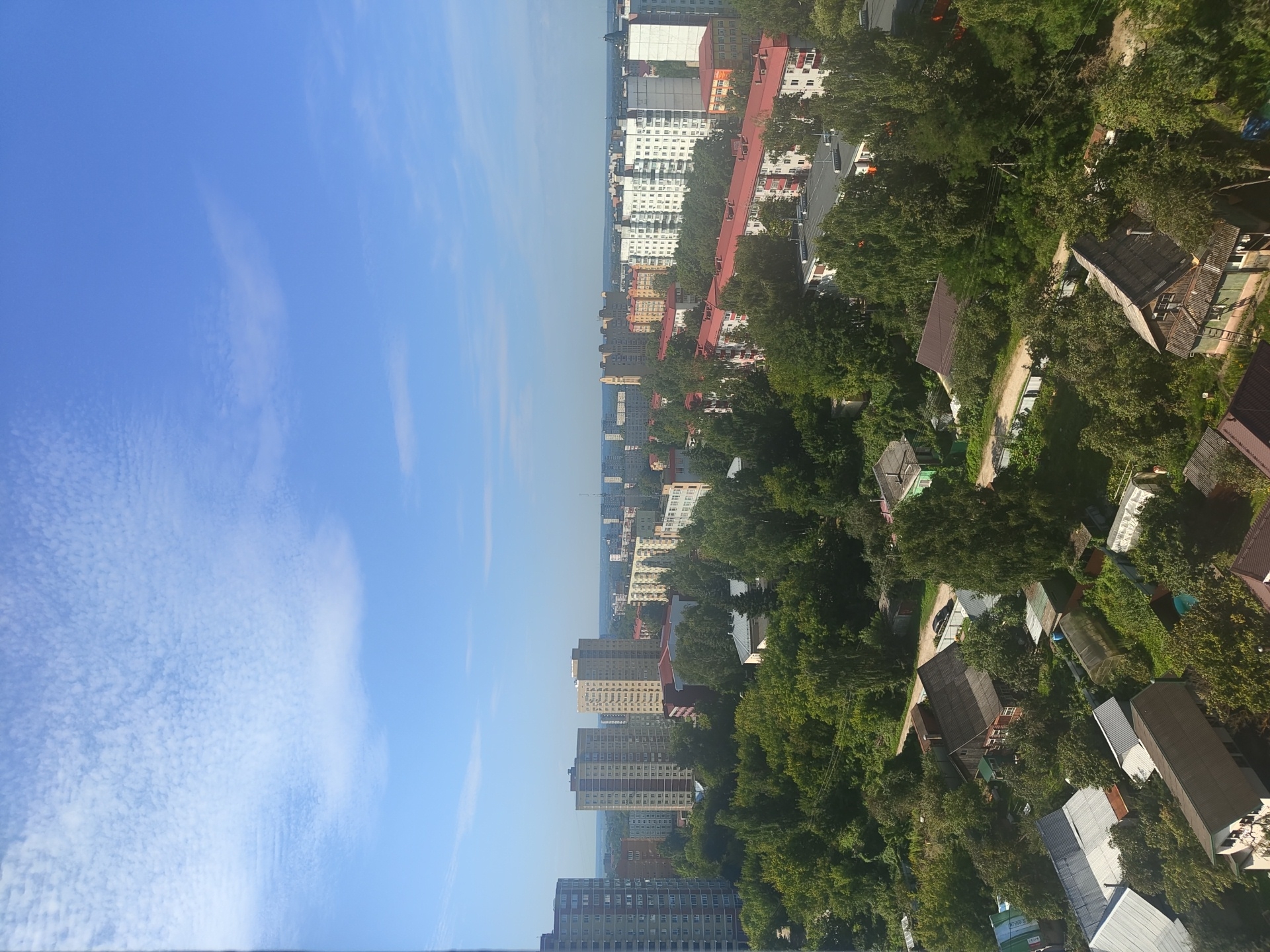 The image is a high-angle, daylight photograph of a residential area in Ramenskoye, Russia. The sky is blue with scattered white clouds. In the background, there are numerous multi-story apartment buildings of varying architectural styles and heights, characteristic of Soviet-era and later urban development. Some of these buildings are light-colored, while others are brick-toned.
In the middle ground and foreground, the landscape transitions to lower-rise buildings, including houses with red and grey roofs, and many trees forming a dense green canopy. There are also some smaller structures that appear to be garages or outbuildings, and possibly greenhouses. A dirt road or path is visible winding through some of the properties in the foreground. The overall impression is of a mixed urban and suburban environment with significant vegetation. There are no people or vehicles explicitly visible in the image.