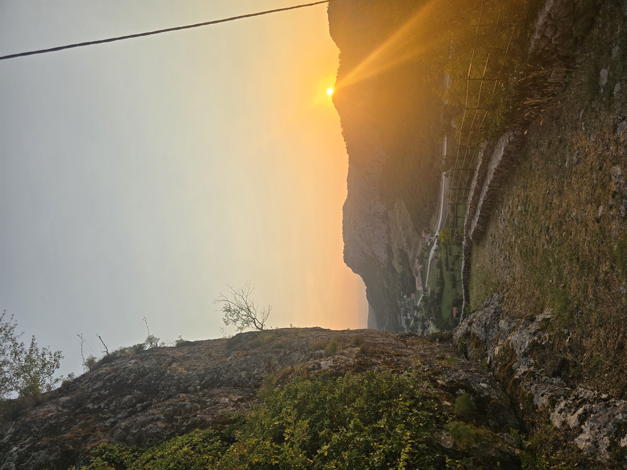 The image depicts a scenic view of the sunset in the vicinity of Gornja Vrbica, Montenegro. The foreground features a rugged cliff face covered in lush green vegetation on the left, and a stone-lined path that curves towards the right. The path is bordered by a low stone wall and a metal railing. In the mid-ground, a valley unfolds with a small village nestled among green hills. A road winds through the valley. The background is dominated by rolling hills and mountains, silhouetted against the bright orange glow of the setting sun. The sun itself is partially obscured by a hilltop, casting long rays of light across the landscape. The sky transitions from a pale blue at the top to a warm orange and yellow hue near the horizon. A thin black wire runs diagonally from the top right corner of the frame into the sky.