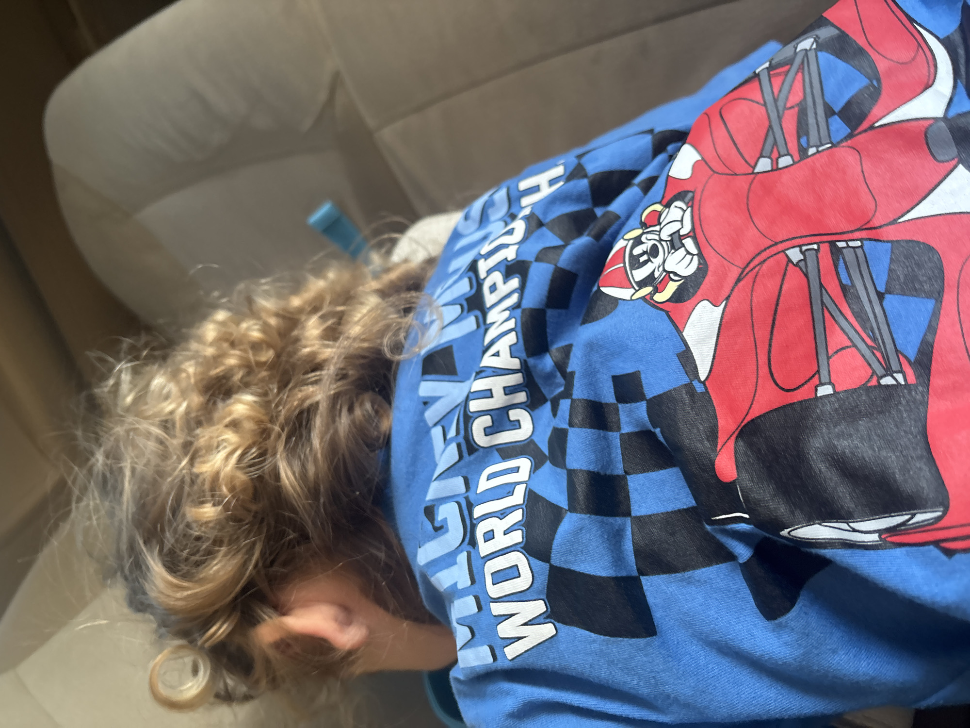 A young child with curly blond hair is sleeping or resting on a tan cushioned seat, possibly in a vehicle. The child is wearing a bright blue t-shirt with a graphic print. The graphic on the back of the t-shirt features a checkered flag pattern and a cartoon image of Mickey Mouse dressed as a race car driver. Mickey Mouse is wearing a red and yellow helmet, driving a red race car with black tires. The shirt also has text that reads "MICKEY MOUSE WORLD CHAMPIONSHIP". The background is blurred, but appears to be the interior of a car or a similar seating area. No specific location in Dubai, United Arab Emirates, can be identified from the image.