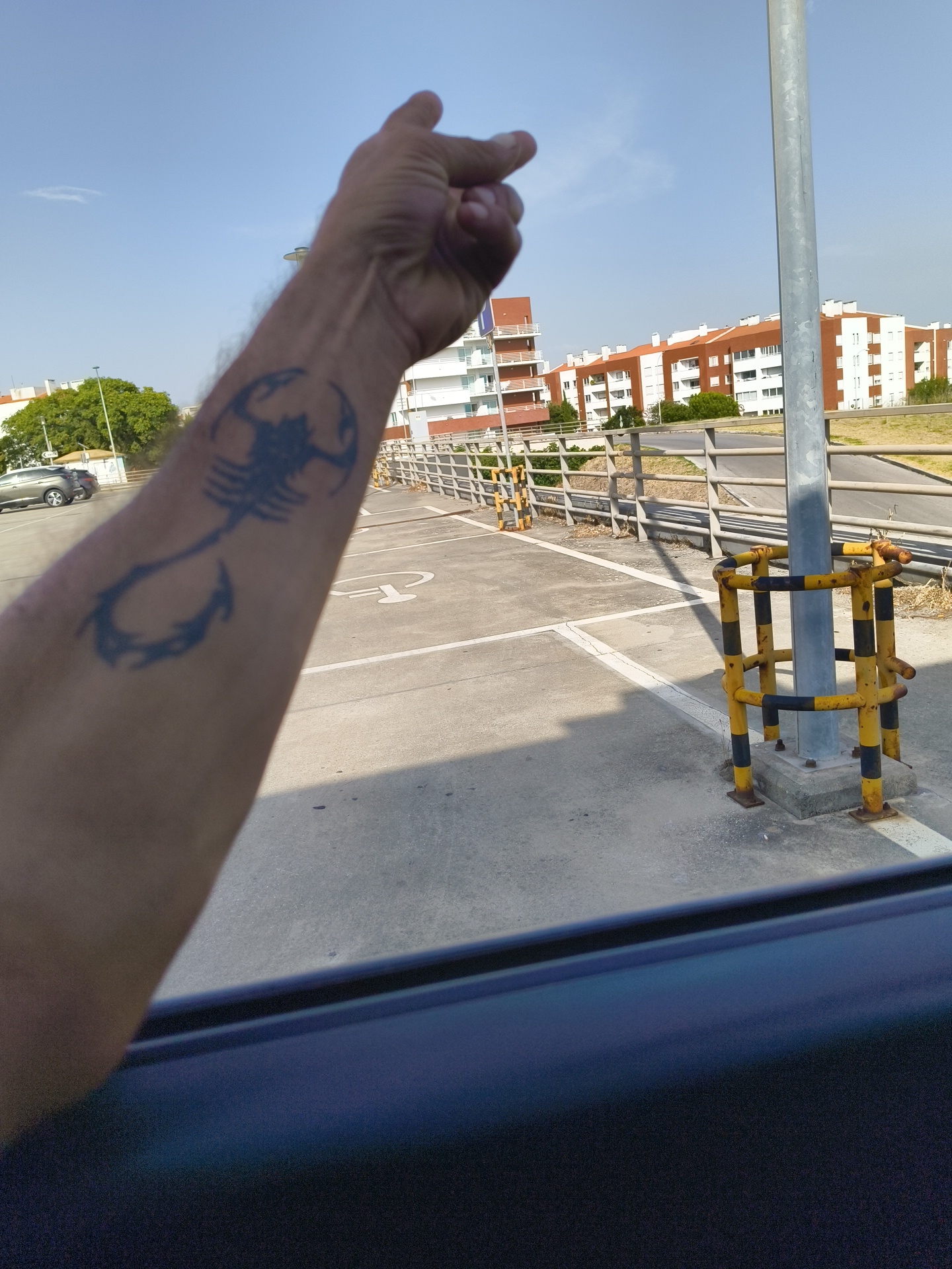 The image shows a close-up of a person's forearm with a scorpion tattoo, with the arm pointing towards the left. The background suggests an outdoor urban environment, possibly a parking lot or street, with buildings and trees visible. The buildings appear to be modern apartment blocks with white and red facades. A metal pole with yellow and black protective barriers surrounds its base. The overall lighting indicates it is daytime with a clear sky. Based on the architectural style and the general environment, this scene could be located in Cascais, Portugal, a coastal city known for its residential architecture and pleasant climate.