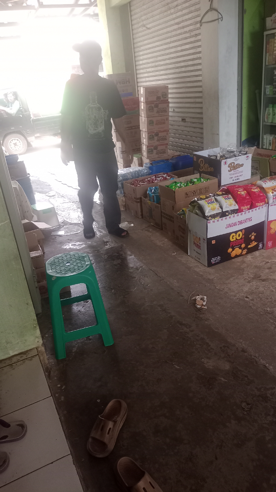 In Cogreg, Indonesia, a person stands in what appears to be a shop or warehouse, partially obscured by bright light from above. They are wearing a dark t-shirt with a graphic and dark pants, standing on a concrete floor that shows signs of wetness. In front of them is a bright green plastic stool. To the right, stacks of cardboard boxes filled with packaged snacks are visible, including some with "Billion" and "GO! PRIME" branding. A roll-up metal door dominates the background. To the left, a truck is partially visible, along with various boxes and shelving. Several pairs of brown sandals are scattered on the floor.