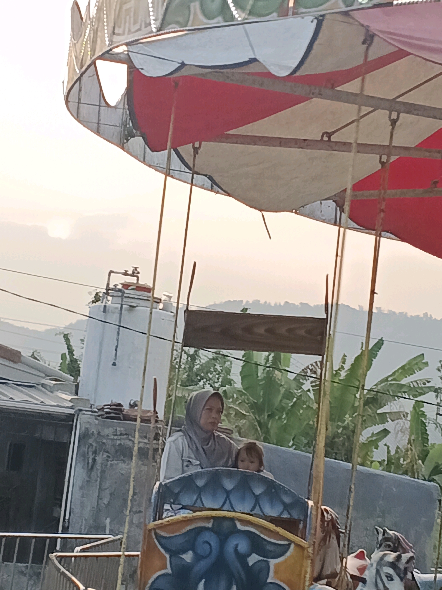 An outdoor shot captures a carousel ride at dusk. The sun is setting, casting a warm glow over a hazy sky. In the foreground, a woman wearing a hijab and a light-colored shirt sits on a carousel horse, holding a young child. The carousel features a colorful canopy and ornate decorations. In the background, residential buildings, a water tank, and lush greenery are visible, suggesting a semi-urban or suburban setting within Sukabumi, Indonesia. The overall atmosphere is peaceful, with the carousel ride appearing to be a local attraction.