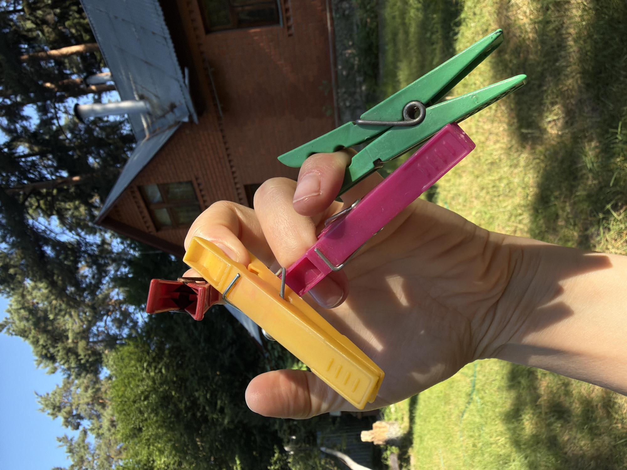 A human hand holds four clothespins: one red, one yellow, one pink, and one green. The red clothespin is small and partially obscured by the yellow one, which is a vibrant yellow. The pink clothespin is also a bright pink, and the green one is a typical grass green. The clothespins are all plastic and have metal springs. The hand is positioned in the foreground, with the clothespins held between the thumb and fingers. In the background, a brick house with a gray, corrugated metal roof is visible. The house has several windows and two chimneys. Lush green trees and a grassy lawn surround the house. The sky is clear and blue. The scene appears to be outdoors on a sunny day. The location is identified as Udelnaya, Russia.