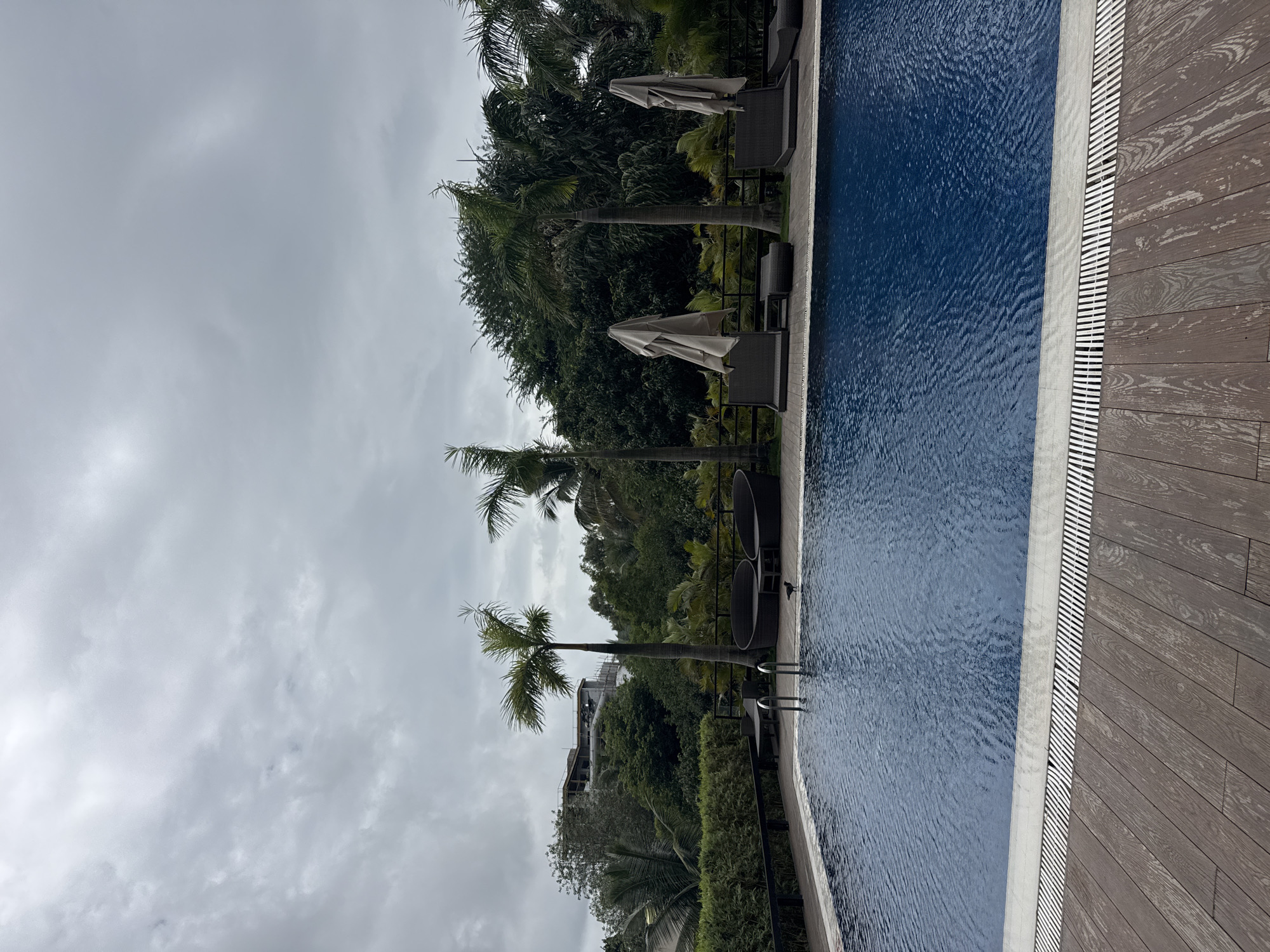 A wide-angle outdoor photograph captures a swimming pool area under a cloudy sky in Pune, India. The pool, filled with deep blue water, occupies the majority of the foreground. A wooden deck with a textured, planked surface surrounds the pool. Several lounge chairs with tan umbrellas are positioned by the pool, suggesting a resort or hotel setting. Palm trees and lush green foliage form a backdrop, with a building under construction visible in the distance. A small black bird stands near the edge of the pool.