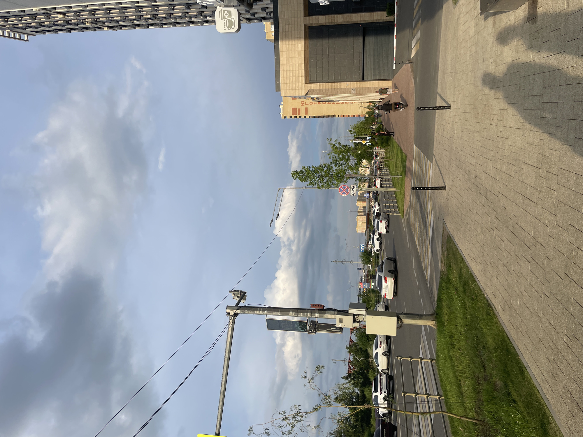 The image depicts a street scene in Stavropol, Russia, during the daytime. The perspective is from a low angle, looking up towards the sky and across a paved sidewalk and a road. On the left, a modern building with a metallic facade rises, featuring rows of parallel lines. To the right of this building is another, taller structure with a beige stone exterior and dark tinted windows. A sign with the word "BRUNCH" and a coffee cup logo is visible on the first building.
The road is lined with parked cars, primarily white sedans, suggesting a busy urban environment. A green grassy area separates the sidewalk from the road. In the distance, more buildings and trees are visible under a partly cloudy sky. The lighting suggests it is either late afternoon or early evening, with long shadows cast across the sidewalk.
There are a few people visible in the scene, walking on the sidewalk or near their vehicles, and one person appears to be riding a scooter or a similar personal mobility device. A traffic light pole with various electronic components and a digital display is a prominent object in the foreground. A red circular sign with a diagonal line through it, possibly indicating "no stopping" or a similar restriction, is also visible. The overall mood is one of everyday urban life.