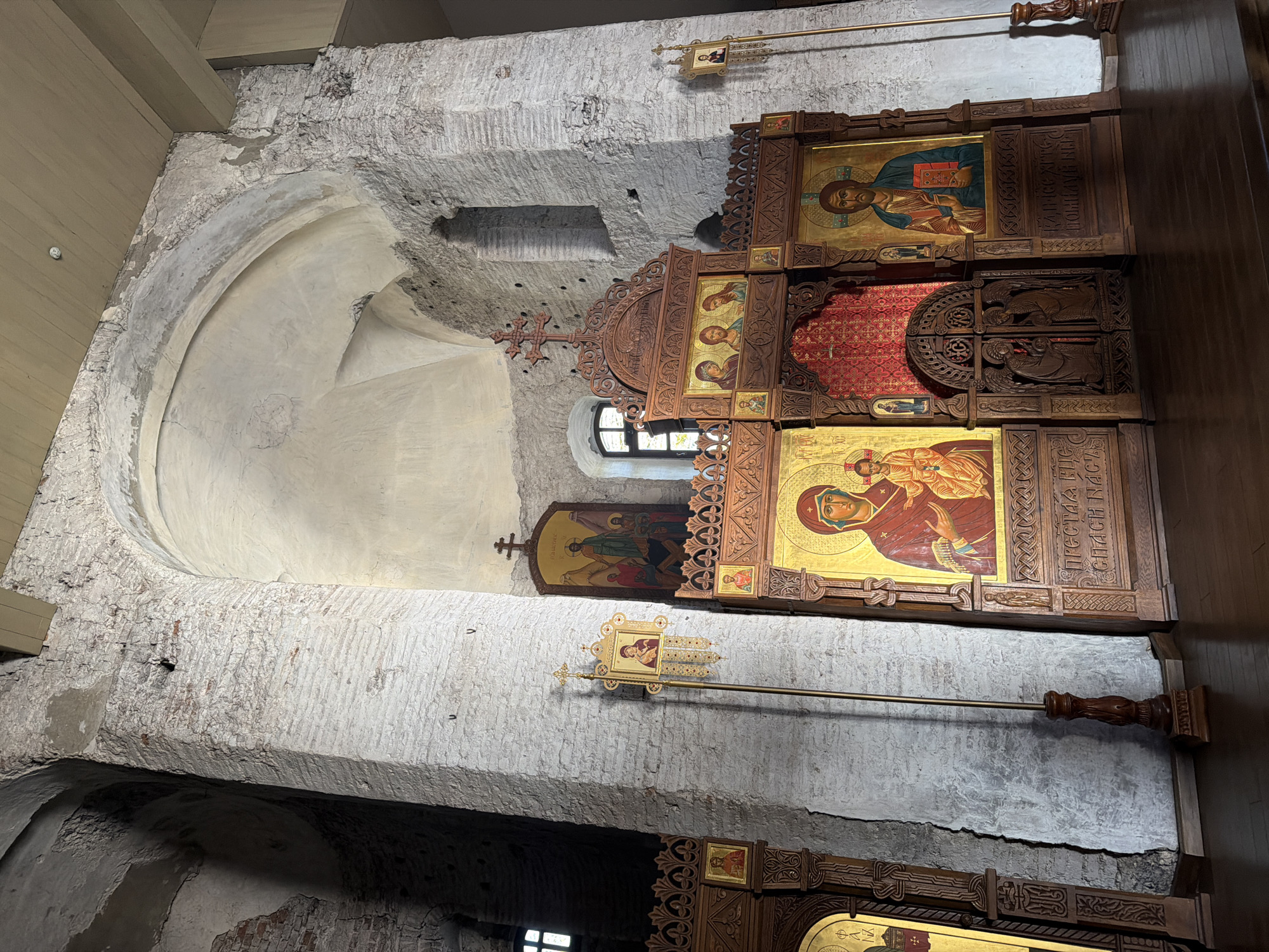 This media file captures the interior of a church or chapel, likely of Eastern Orthodox tradition, in Hrodna, Belarus. The focal point is a multi-tiered wooden iconostasis, intricately carved and adorned with golden-framed icons.
Prominently displayed on the iconostasis are several icons. The top right icon depicts Jesus Christ as Pantocrator, holding a book, with visible Old Church Slavonic text that includes "ПРЕСТЛА" and "СЪНАВЫ". Below this is another icon of the Theotokos (Virgin Mary) holding the infant Jesus, also featuring Old Church Slavonic script, such as "ПРЕСТАА ВЛАСДИЧ" and "СПАСЫ". Further down, a partial view of another icon is visible. To the left, integrated into an arched wall section, is an icon depicting a group of figures, possibly saints or a biblical scene.
The surrounding architecture features rough, white-washed stone or brick walls, showing signs of age and texture. Arched alcoves and a small, arched window allowing natural light are visible. The ceiling above appears to be constructed from modern, light-colored wooden beams, contrasting with the older stonework. Leaning against the wall or iconostasis is a long, golden liturgical staff or banner, topped with a small icon depicting the Virgin Mary and child.
The scene conveys a sense of reverence and historical depth, characteristic of a religious space. No direct activity is taking place, but the setup indicates readiness for worship.