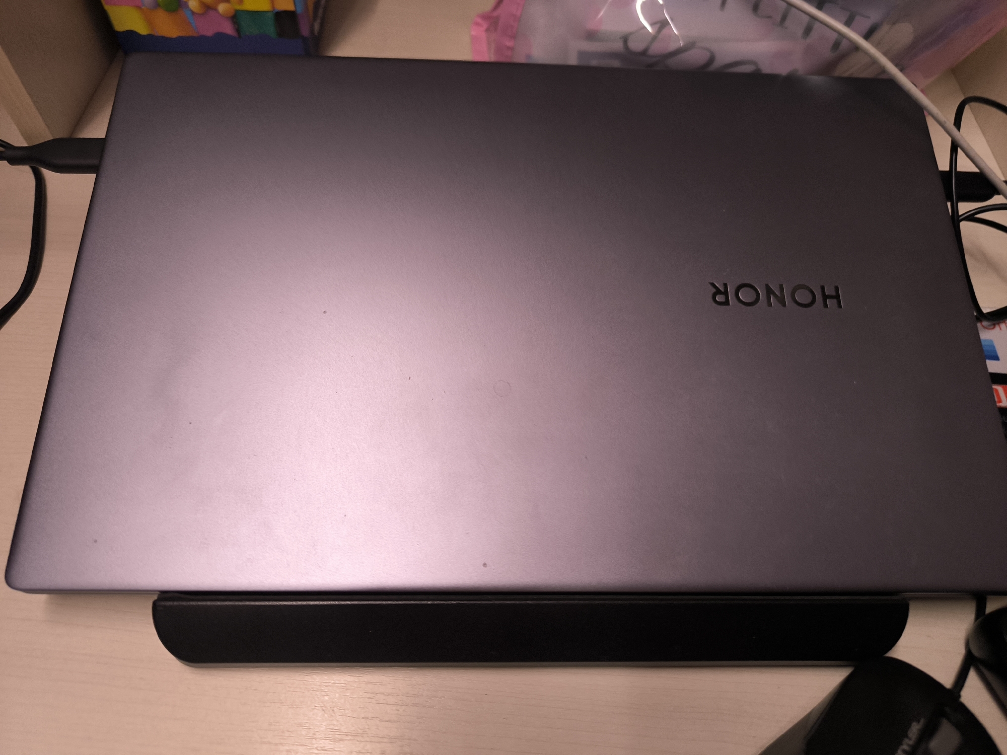 A closed, dark gray laptop with the "HONOR" logo embossed on its lid sits on a black stand. The laptop is placed on a light-colored wooden desk. To the left of the laptop, a black cable is plugged into its side. In the background, a colorful object with a mosaic-like pattern is partially visible, along with a pink plastic bag containing various items, possibly office supplies. The overall scene suggests a home or office environment. The location is not specified in the image.