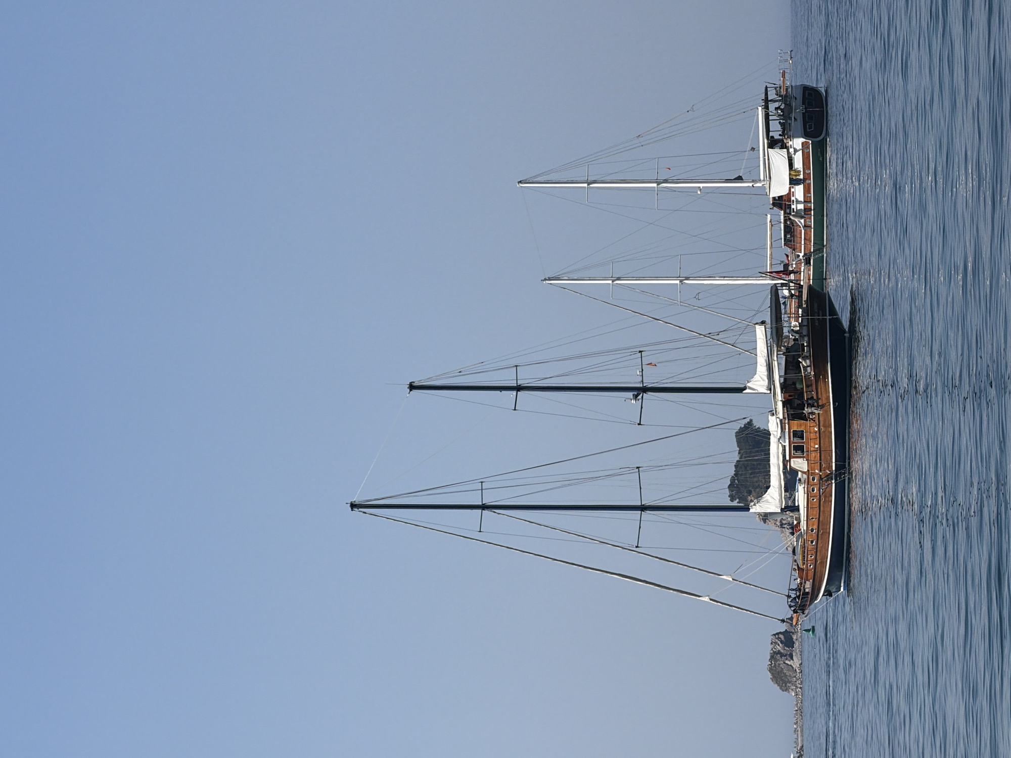 This image features a side view of a wooden sailing vessel with multiple masts anchored in clear, blue water. The ship is oriented vertically in the frame, with its hull running from the bottom to the top of the image. Three prominent masts rise from the deck, supporting a complex rigging system, though no sails are deployed. The deck itself appears to be made of wood, with several cabins and windows visible. A few people can be seen on the deck, engaged in what appears to be leisurely activity.
The water is calm and rippled, reflecting the clear, light blue sky. The scene suggests a bright, sunny day, likely in the morning or late afternoon given the quality of light. In the background, rocky islands or a coastline can be seen, adding to the maritime setting. The overall mood is peaceful and serene, highlighting the beauty of the sailing vessel and its natural surroundings. There is no visible text in the image.