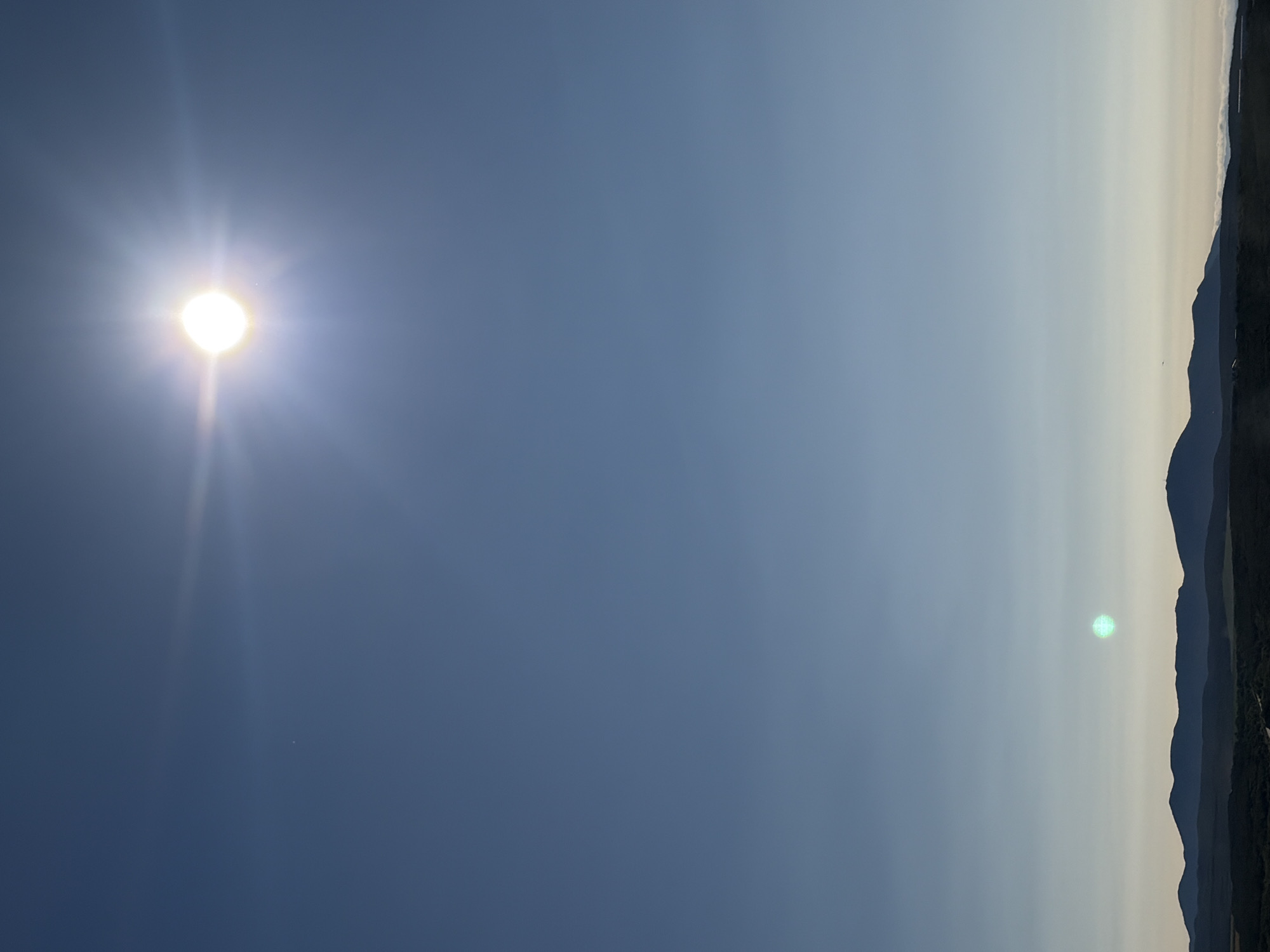 The image captures a wide-angle view of a vast, clear blue sky with the sun prominently positioned in the upper right quadrant, emitting a bright glare. A small, faint light, possibly a distant celestial body or artifact, is visible in the upper left portion of the sky. In the lower half of the image, a silhouette of a mountain range stretches across the horizon, with dark, undulating peaks against a lighter sky. The foreground depicts a dark landscape, likely covered in vegetation or trees, with a hint of a road or pathway winding through it. A faint green light, possibly lens flare or atmospheric phenomenon, is present in the lower middle section of the image, above the mountains. The overall scene suggests an outdoor environment, likely a scenic overlook or high vantage point, with no human presence or specific urban features identifiable. The image does not contain any elements that would allow for identification of the location in Korsakov, Russia.