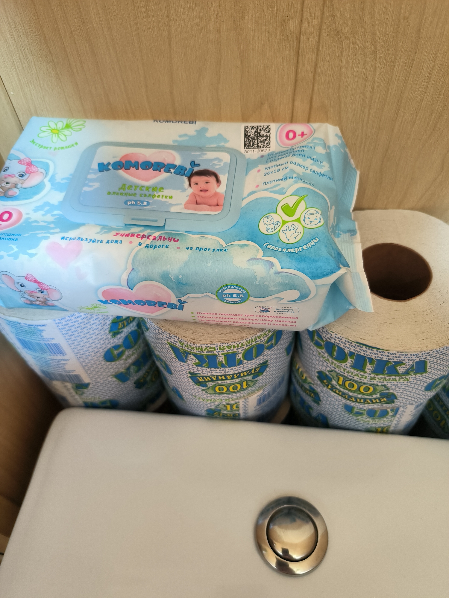 The image shows a close-up of a package of KOMOREBI baby wipes partially covering several rolls of toilet paper. The wipes package is blue and white with cartoonish imagery of a baby and a bunny. The text on the package is in Russian. The toilet paper rolls are white and also have Russian text and imagery printed on their outer wrapping, which appears to be a repeating pattern of "100" and a blue and green design. The background is a light brown wooden surface. The scene appears to be an indoor domestic setting, possibly a bathroom, given the presence of toilet paper. There are no people visible in the image.