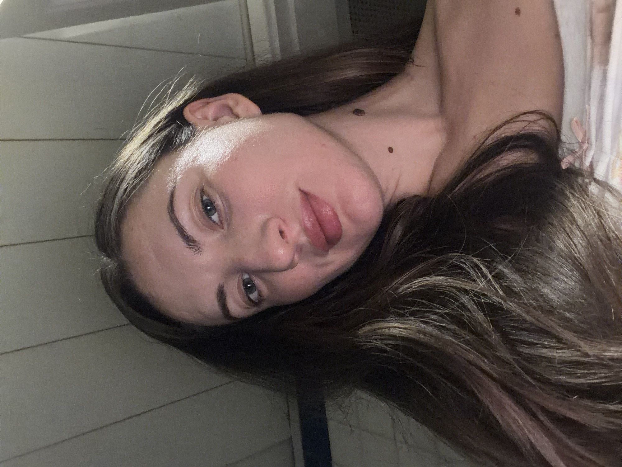 The image is a close-up, low-angle selfie of a woman's face and upper chest. She has long, dark brown hair with lighter streaks, which frames her face. Her skin appears fair and luminous. She has light blue eyes, full lips, and a prominent mole on her neck below her jawline, as well as another smaller mole on her collarbone. The background appears to be an indoor setting with horizontal wood paneling on the ceiling and a dark horizontal band which might be a shadow or an object. There is no indication of the city of Sharm El Sheikh, Egypt, or any other specific location. The subject is wearing a light-colored top with a faint floral pattern. The lighting suggests it is evening or night.