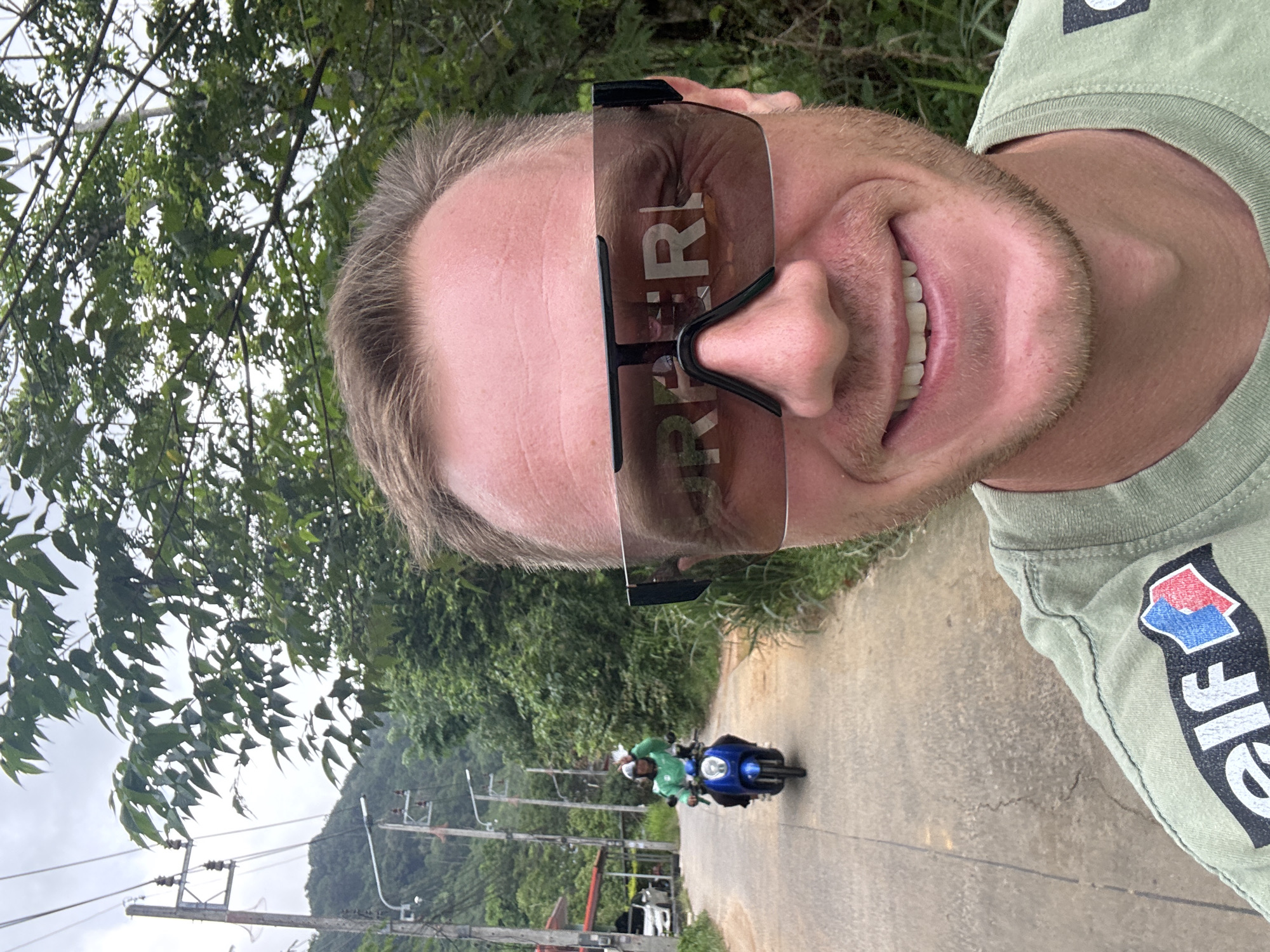 In Rawai, Thailand, a man with short blonde hair and a beard takes a selfie outdoors on a paved road lined with trees and electrical poles. He is wearing a light green t-shirt with an "elf" logo on the left sleeve and a dark-rimmed wrap-around sunglasses. His sunglasses reflect the word "BURBERRY" in large white letters. In the background, a person wearing a green shirt rides a blue scooter. The road leads uphill and curves to the left, disappearing behind foliage. The sky is overcast.