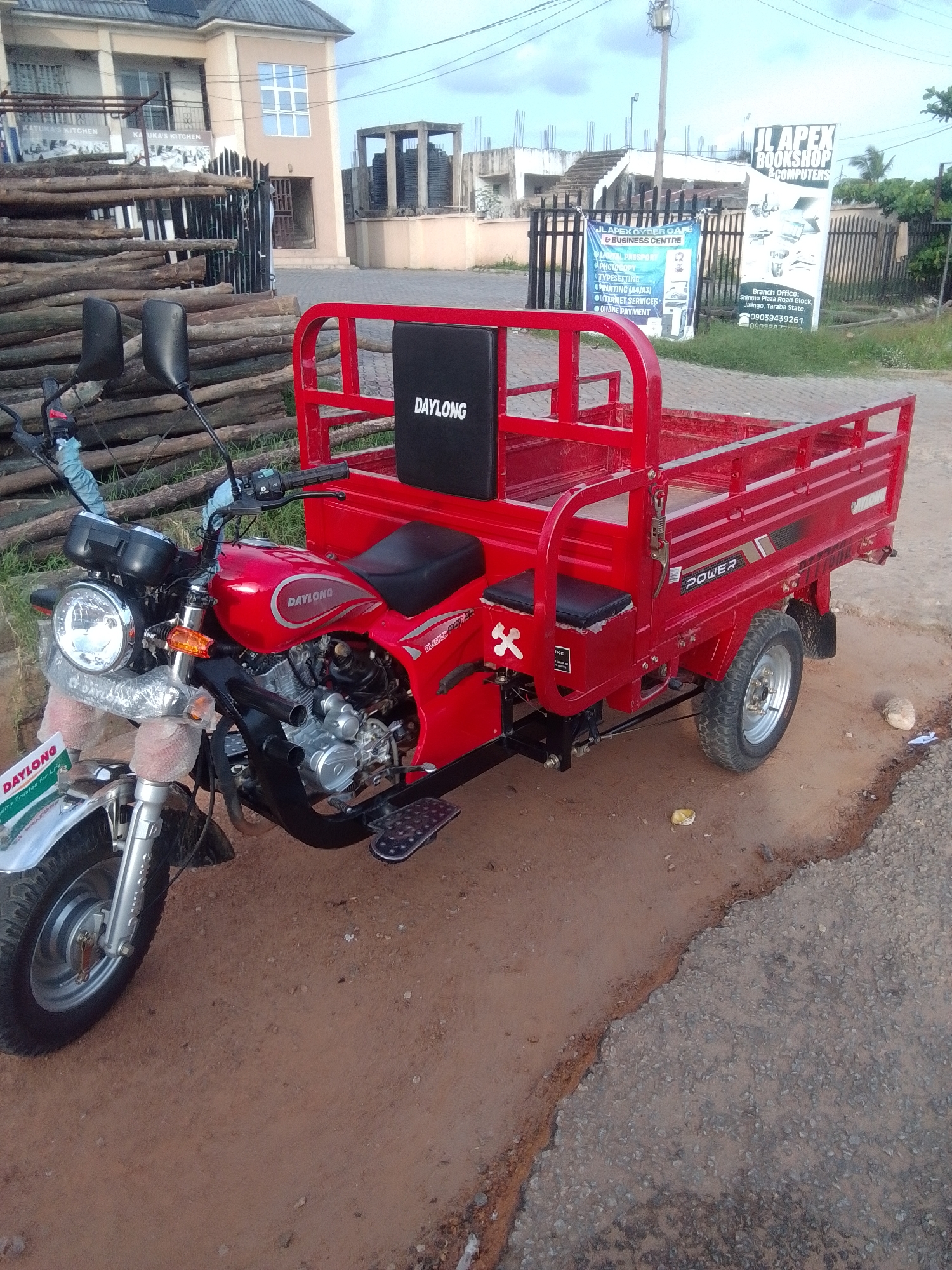 A red, three-wheeled vehicle, possibly a cargo tricycle, is parked on a dirt and paved road. The vehicle has a front motorcycle-like section with a headlight and handlebars, and a rear cargo bed with red metal rails. The seat of the vehicle has a black cushion with the brand name "DAYLONG" printed on it in white. There is also a "POWER" decal on the side of the cargo bed. The background shows a building with multiple windows, a fence, stacks of wooden logs, and a street sign that reads "JL APEX BOOKSHOP & COMPUTERS" with an address in Jalingo, Nigeria. The sky is overcast.