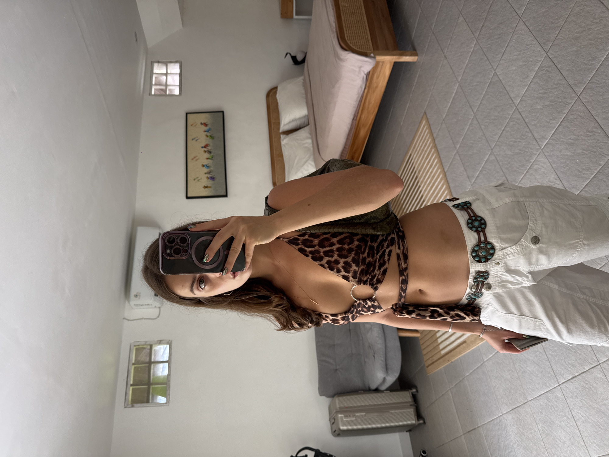 A young woman stands in a bedroom, taking a mirror selfie with her phone. She is wearing a leopard-print halter top with metal ring details, white cargo pants, and a decorative belt with turquoise accents. The room has light grey tiled floors, white walls, a bed with a pink comforter, a grey sofa, and a suitcase. A small framed artwork hangs on the wall. The overall impression is a casual, possibly vacation setting. The image is taken indoors, likely in a private residence.