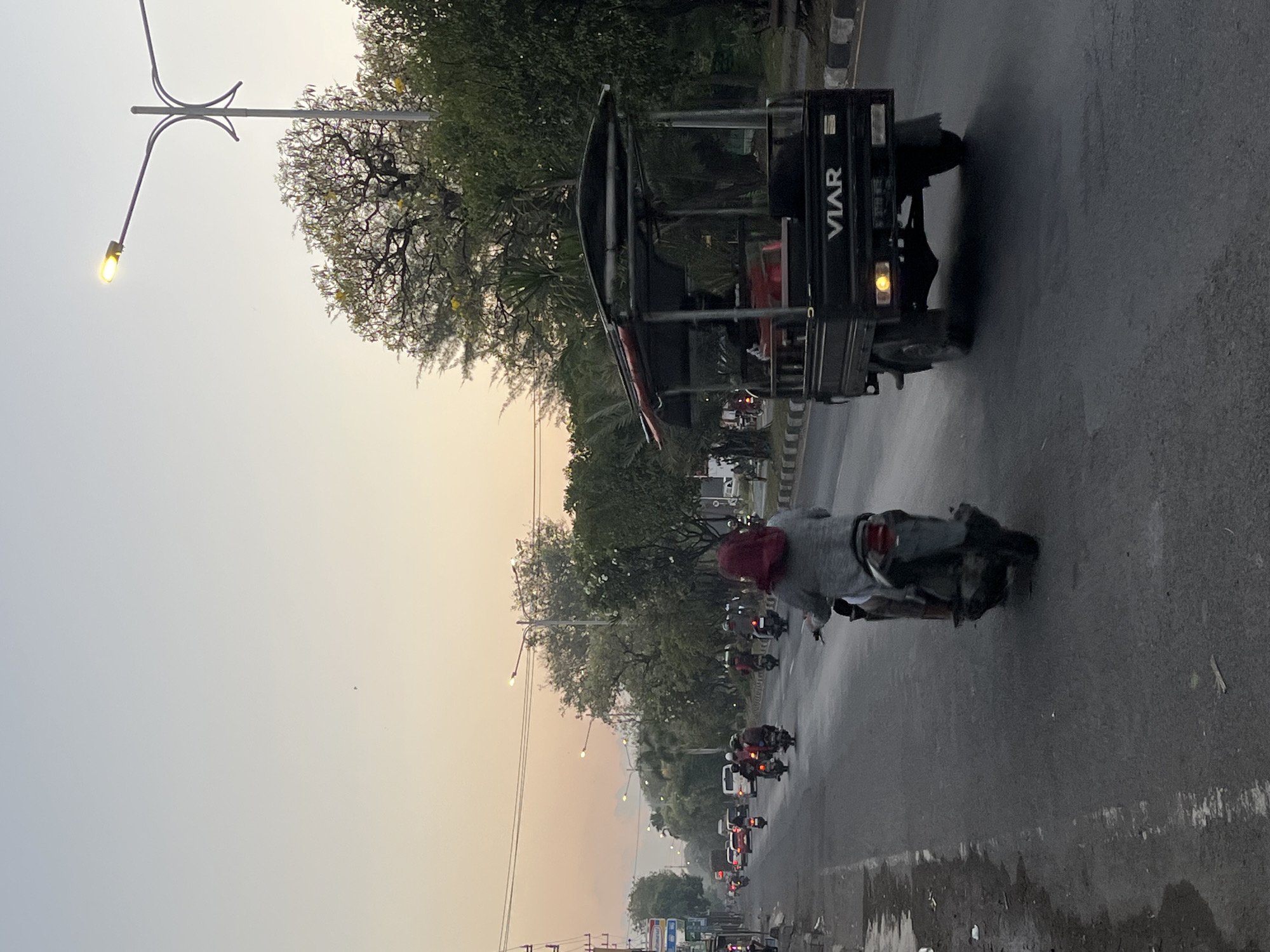 The image depicts a street scene in Ngaban, Indonesia, likely during the late afternoon or early evening, as indicated by the soft, warm light and the illuminated street lamp. The street is paved and appears wet, possibly from recent rain. A variety of vehicles are present, with motorcycles being the most numerous. Several individuals are riding motorcycles, some wearing headscarves. A three-wheeled vehicle, identified by the "VIAR" logo on its rear, is also visible, carrying passengers under a canopy. The roadside is lined with trees, some with visible yellow flowers, and there are buildings and streetlights along the way. The overall atmosphere suggests a typical urban environment with moderate traffic.