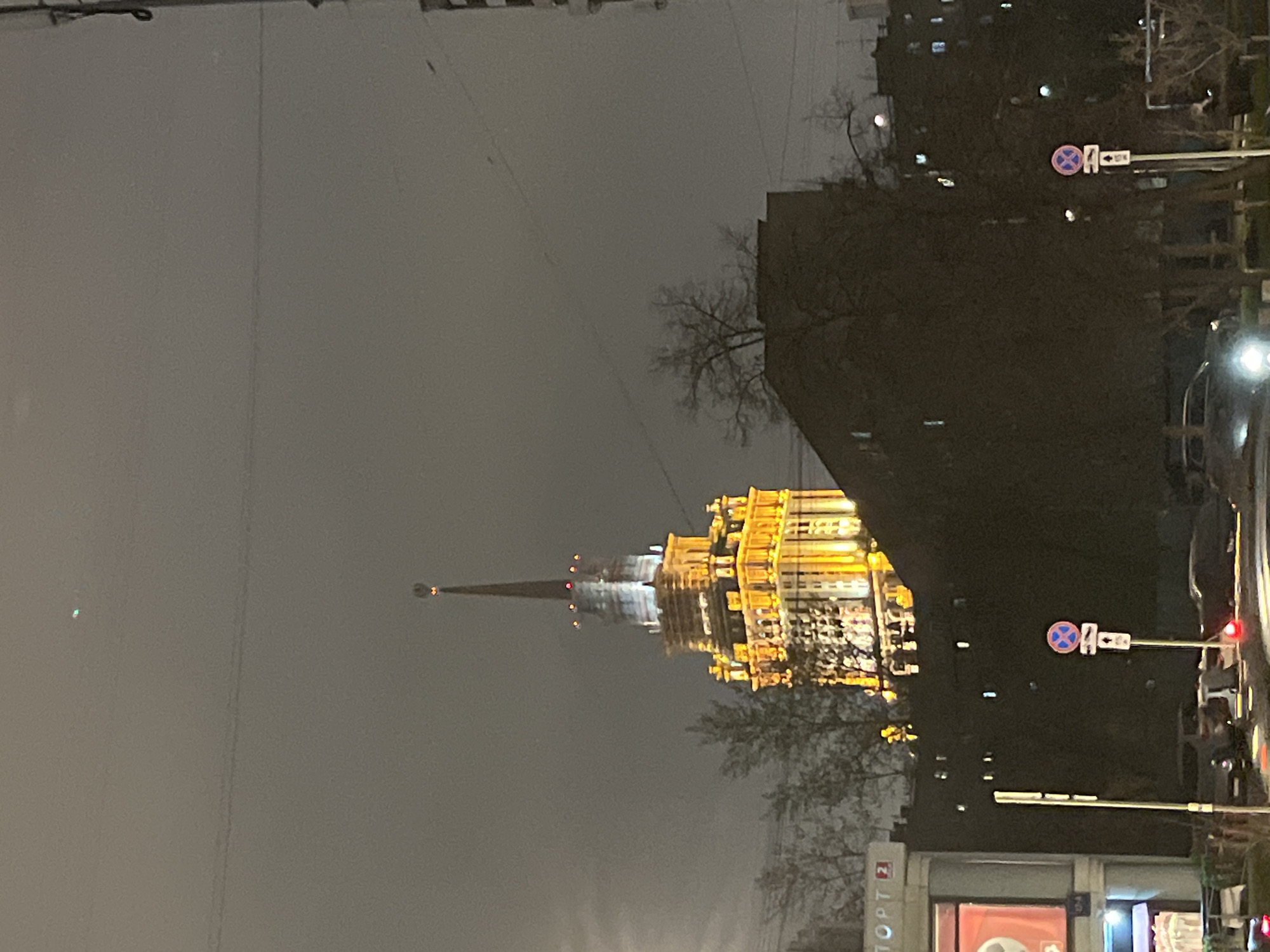 The image shows a night scene in Moscow, Russia, featuring one of the "Seven Sisters" skyscrapers, likely the Hotel Ukraina (Radisson Collection Hotel). The building is illuminated with golden lights, contrasting with the dark, overcast sky. A tall spire extends from the top of the building. In the foreground, there are trees and buildings, including a sign that reads "Спорт" (Sport) in Russian. A "no stopping" traffic sign is visible on the right side of the frame. A street or road runs along the right edge of the image. Overall, the scene depicts a cityscape at night, with a prominent architectural landmark as the focal point.