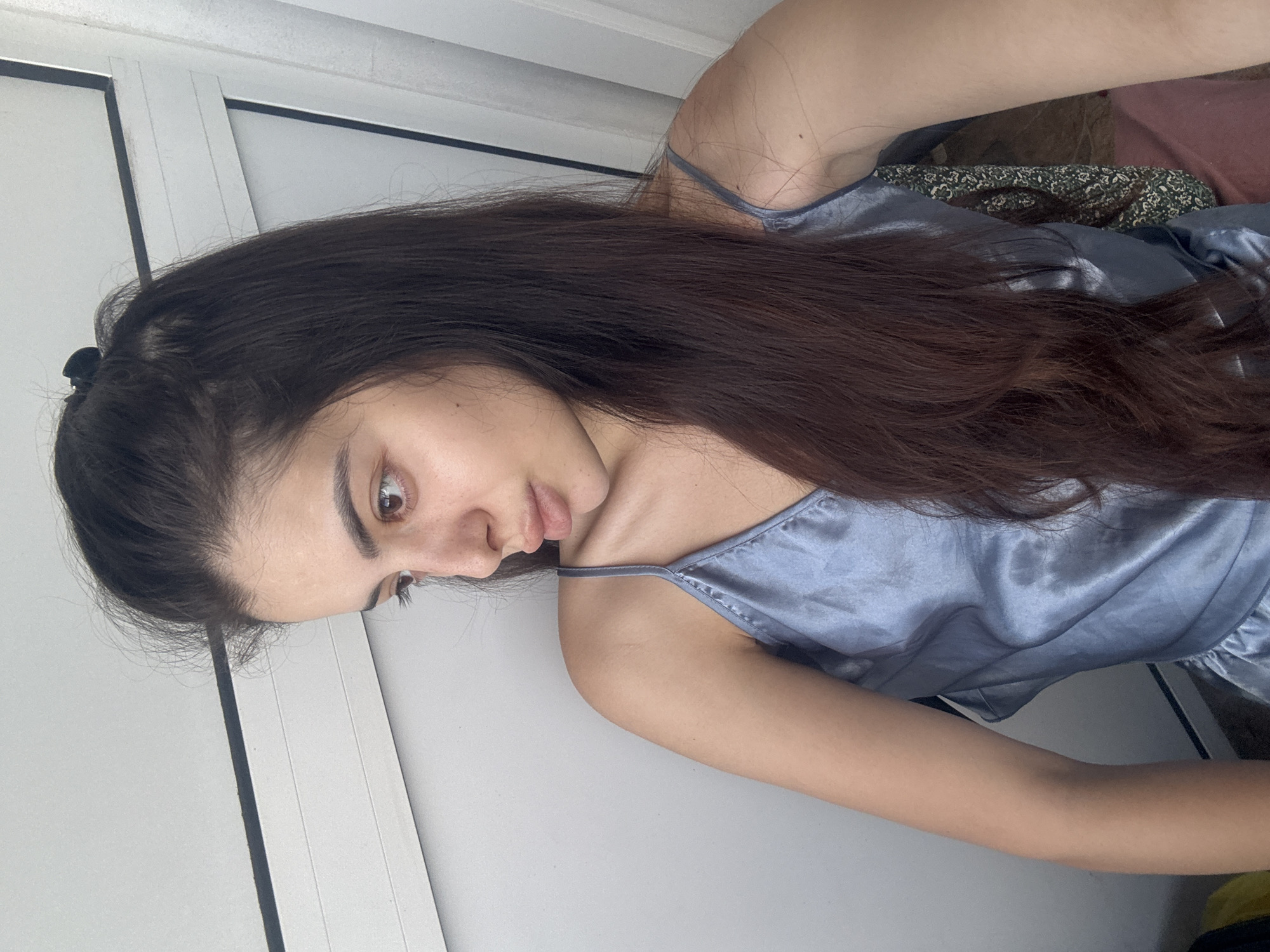 A young woman is depicted in a close-up, diagonally oriented portrait, with the image rotated 90 degrees clockwise. Her head is in the upper left part of the frame, and her body extends towards the lower right. She has long, dark brown hair, with some strands pulled back by a dark hair clip on the crown of her head, while the majority flows over her left shoulder and chest. She is wearing a light blue or grey satin camisole top with thin spaghetti straps. Her skin is fair, with visible freckles across her nose and upper cheeks. Her light brown eyes are open, and she is gazing downwards and slightly to her left, exhibiting a neutral or slightly pensive expression. The background is predominantly a plain white surface, likely a wall or door panels, featuring faint dark lines. To the far right, partially obscured by her hair, there are hints of a dark green patterned fabric and a reddish-brown material. The lighting is bright and even, indicating an indoor setting, likely during daytime. There is no visible text in the image. The scene is set in Plovdiv, Bulgaria.