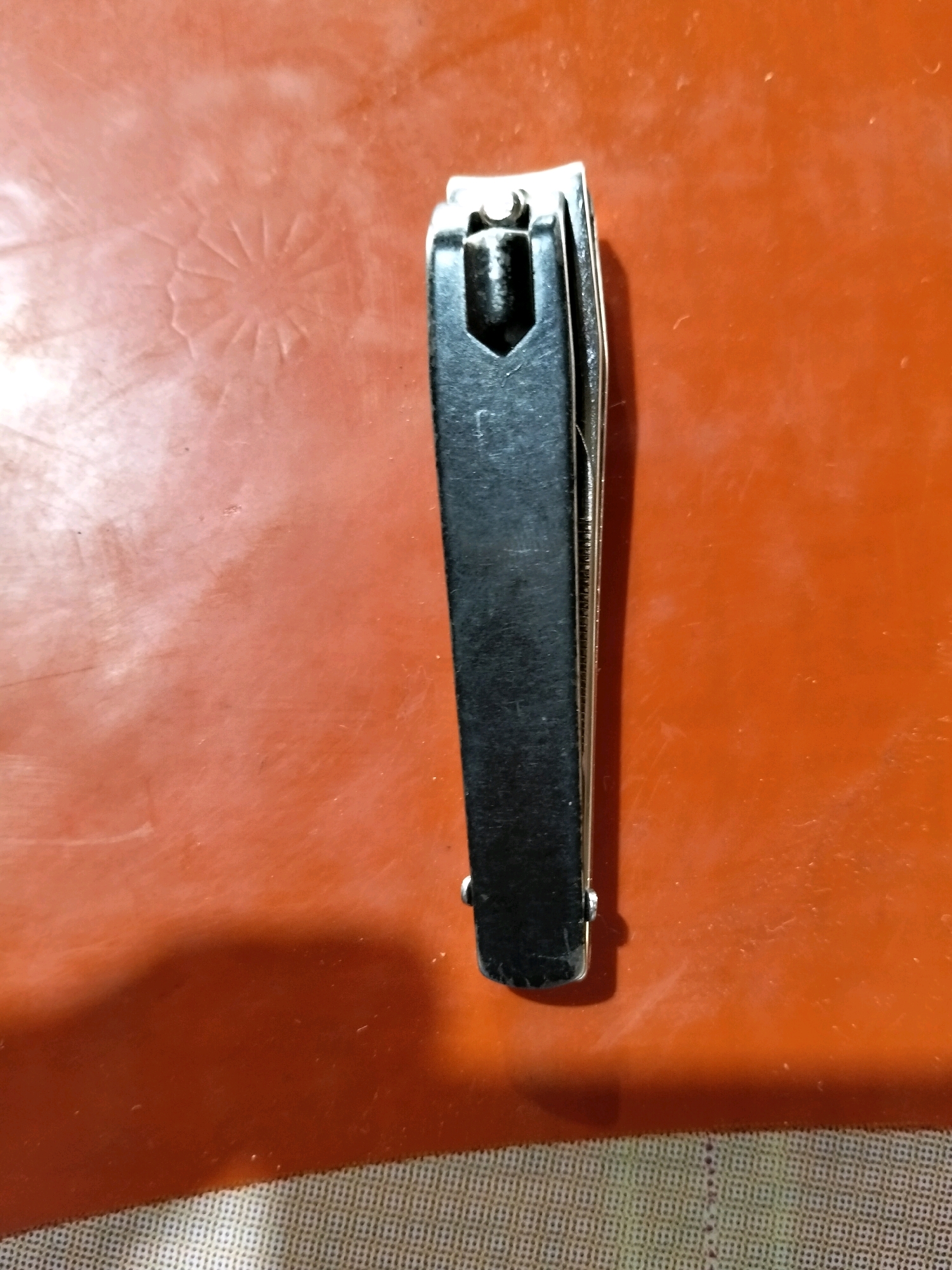 A black nail clipper rests on a textured orange surface, with a faint circular embossed pattern visible on the left side of the surface. The nail clipper is positioned vertically, with its top end slightly angled. The background appears to be a flat surface with some subtle texture and a shadow cast to the lower left. The image is a close-up, top-down view of the nail clipper.