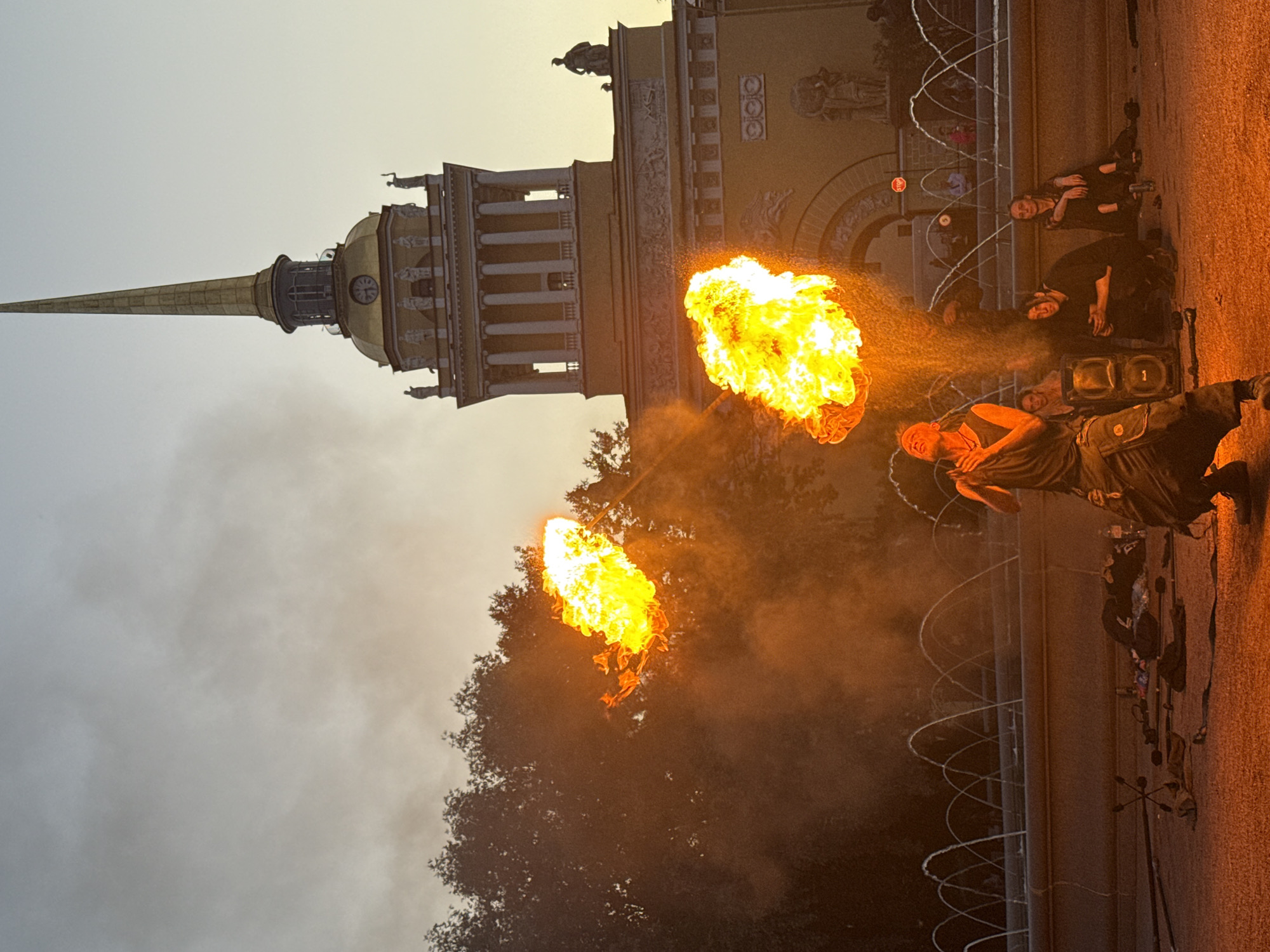 The image captures a fire performance at dusk in Palace Square, Saint Petersburg, Russia. The central figure, a male fire performer, is in the act of expelling a large plume of fire from his mouth, creating a dramatic visual effect. He is dressed in dark, loose-fitting clothing, appearing to be in mid-performance. To his left, another blast of fire erupts, illuminating the surrounding trees and smoke. In the background, the Admiralty Building, a prominent landmark in Saint Petersburg, stands tall with its distinctive golden spire. Fountains are visible in front of the building, adding to the ambiance of the square. Several spectators are seated or standing in the middle ground, observing the performance. Objects related to the performance, such as fire-breathing props and a speaker, are scattered on the ground in the foreground. The sky is a muted gradient of orange and grey, indicating the transition from day to evening.