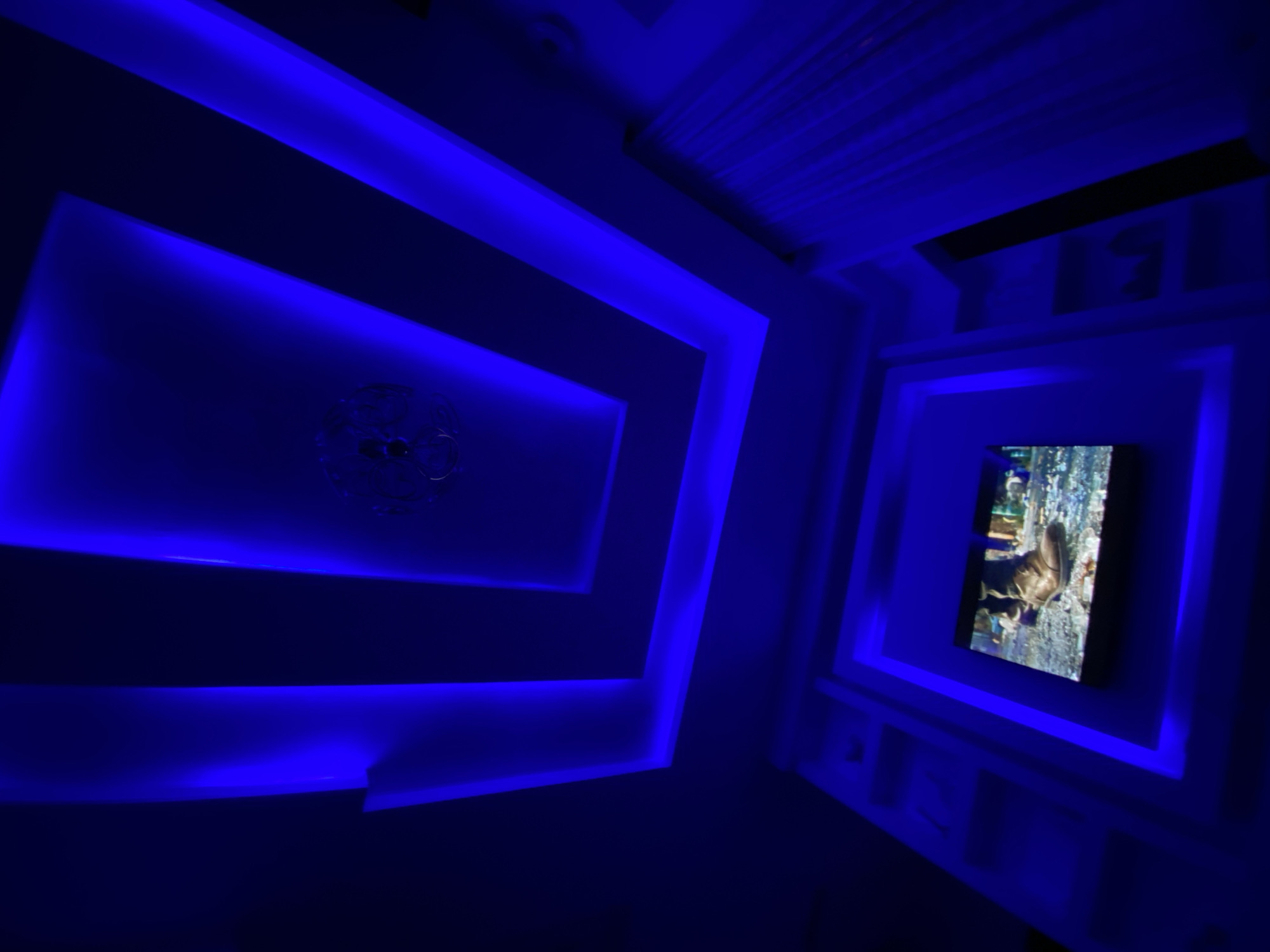 The image depicts a room interior with blue lighting fixtures. The design incorporates rectangular, recessed ceilings with integrated blue LED strips, giving a modern and stylized aesthetic. A chandelier is visible within the larger rectangular design on the ceiling. In the right section of the image, a television screen displays an image, mounted within a similar rectangular recess, also illuminated with blue LEDs. The location, Lagos, Nigeria, is not discernible from the image alone. The description is limited to the observable elements within the photograph.